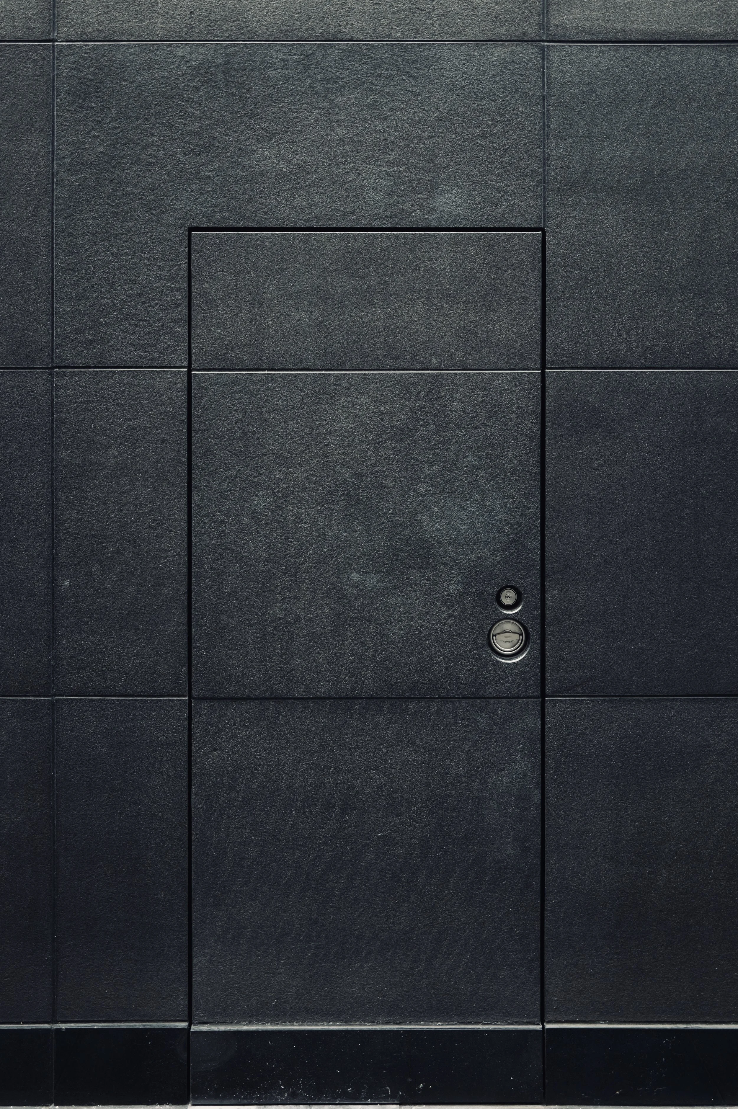 A flush rectangular door with two round fittings set into a dark wall of large stone panels.