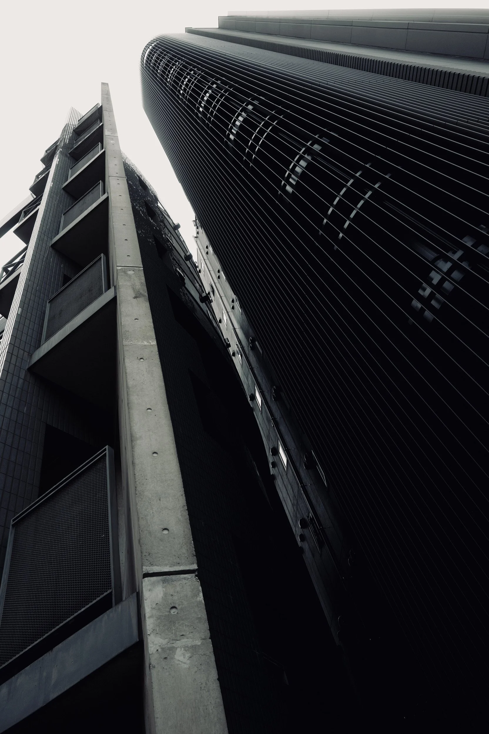 A narrow wedge of sky between a concrete building and a curved facade lined with dark vertical fins.
