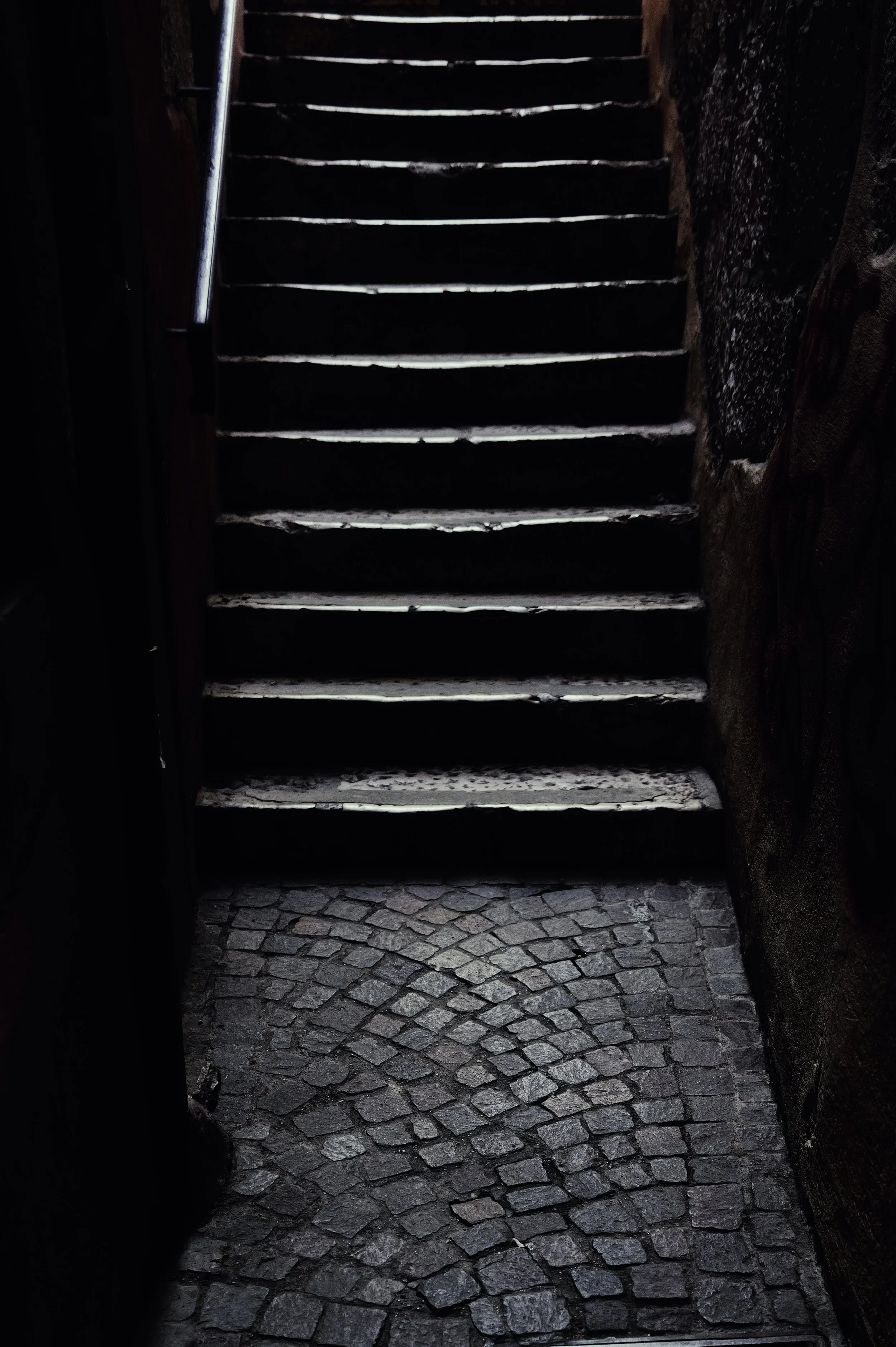 Atmospheric photography of a wooden staircase between stone walls leading toward natural light. Vertical structure.