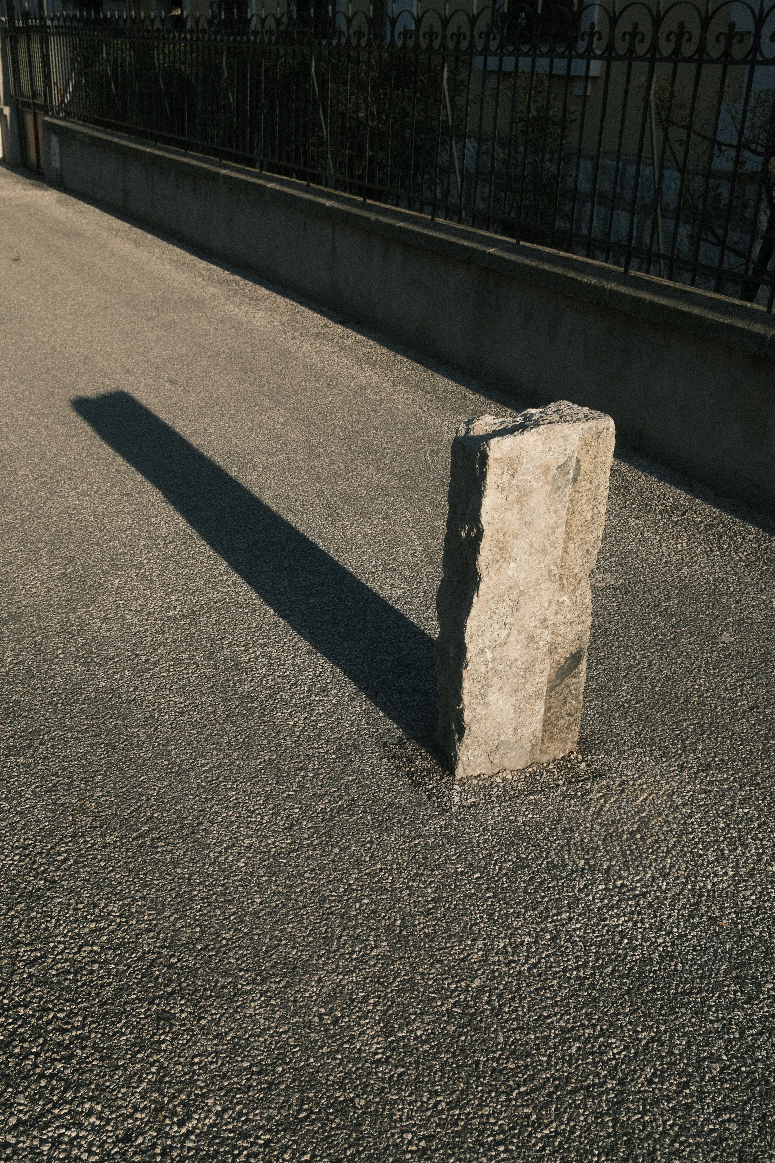 Minimalist street detail featuring a stone post and long dramatic shadow on a sidewalk. Focus on light and silence.