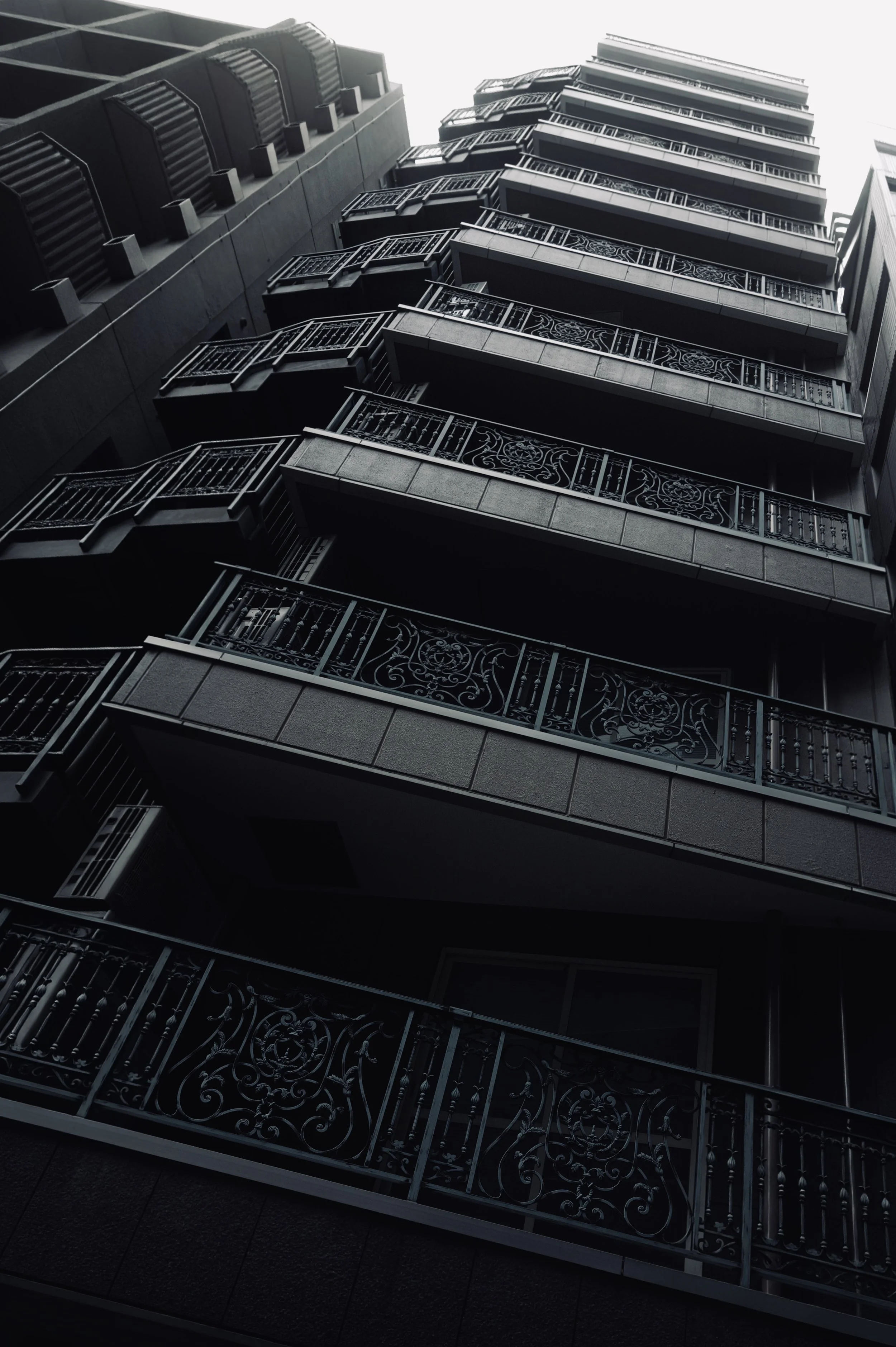 A dark building seen from below, with stacked projecting balconies and patterned metal railings beneath a pale sky.