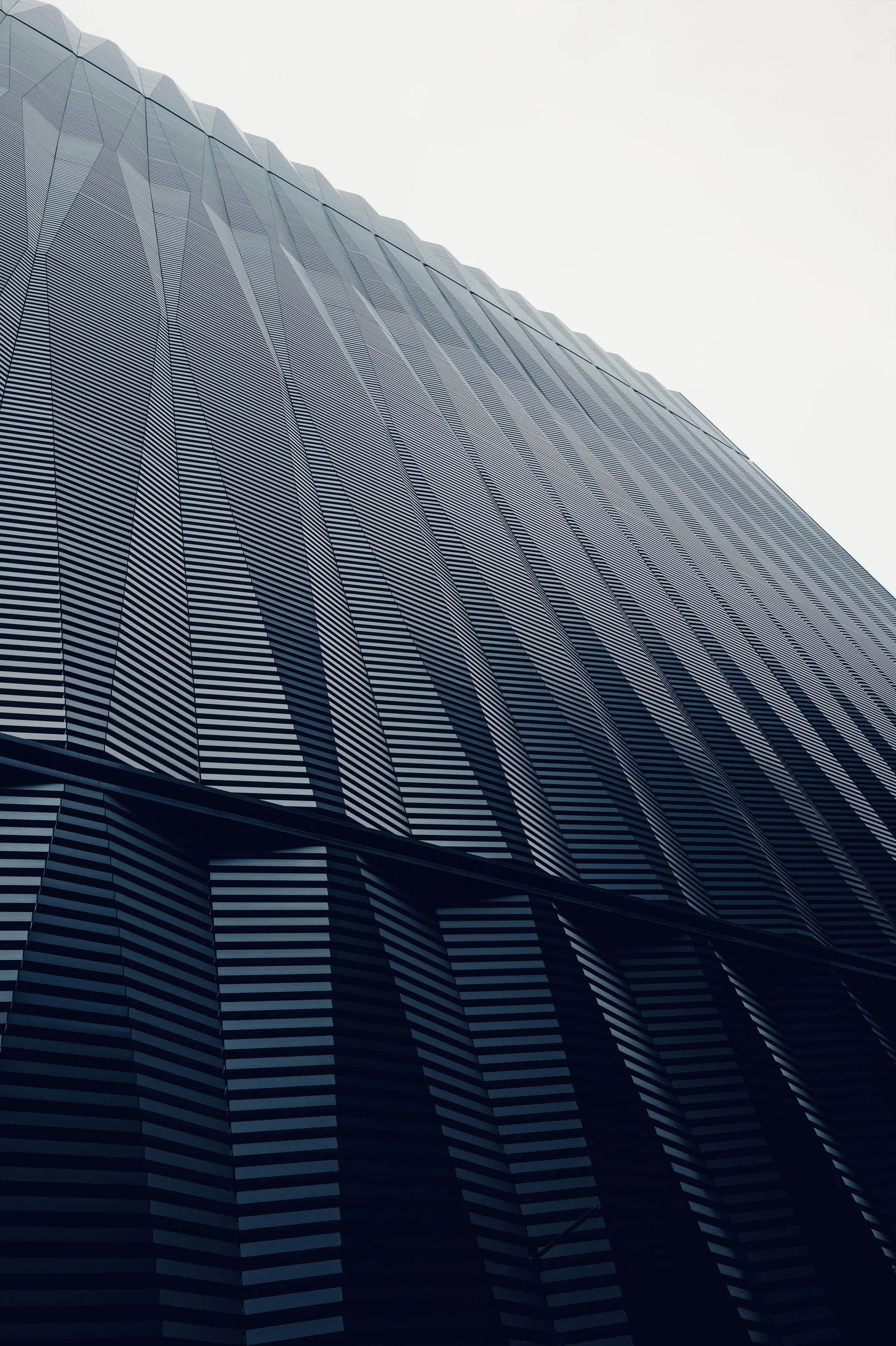 A ribbed metal facade rising diagonally into a pale sky, with blue-grey shadows.