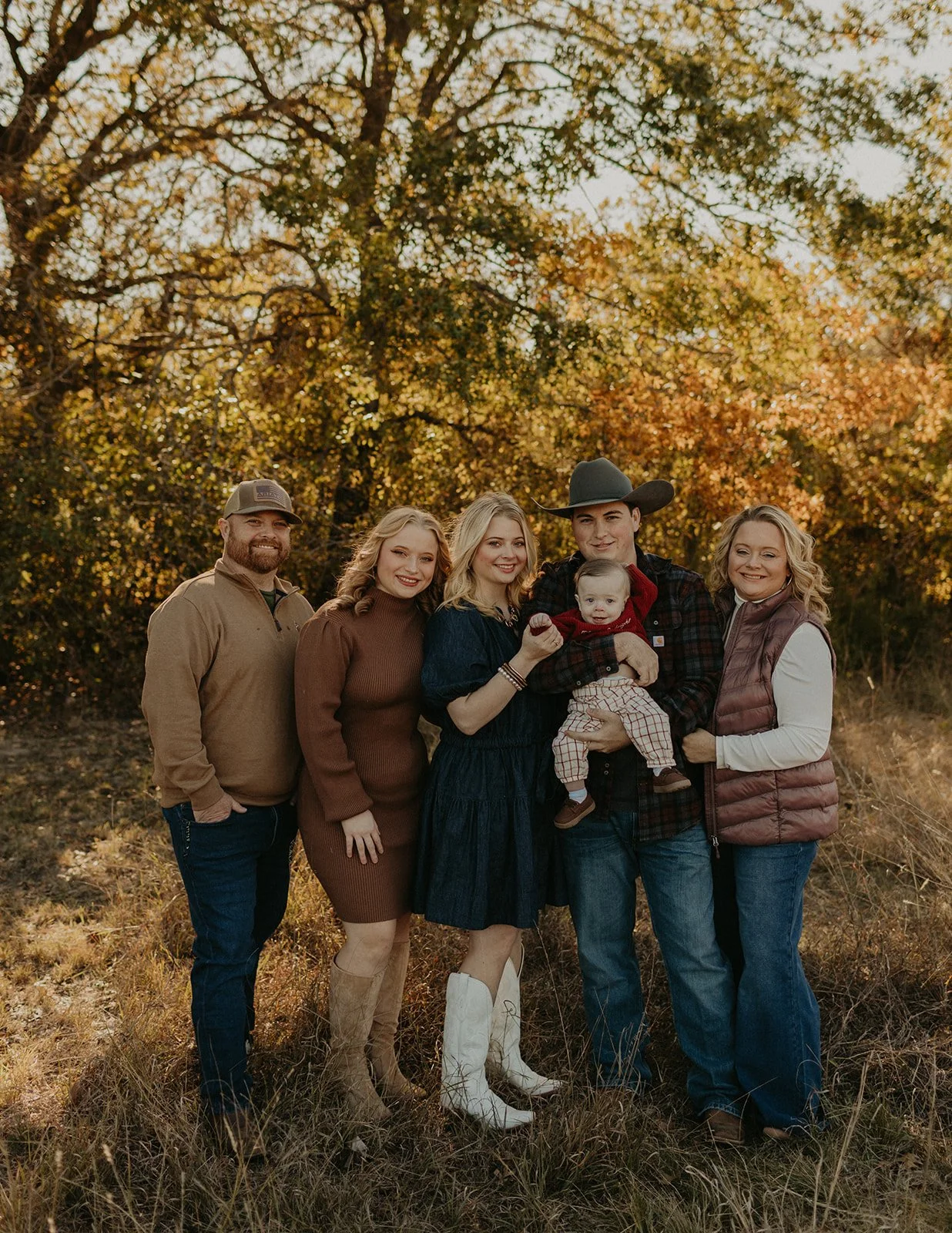 A family of six standing outdoors in front of autumn trees, smiling. The group includes three women, one man, a young boy, and a baby, dressed in fall clothing, with warm tones and casual attire.