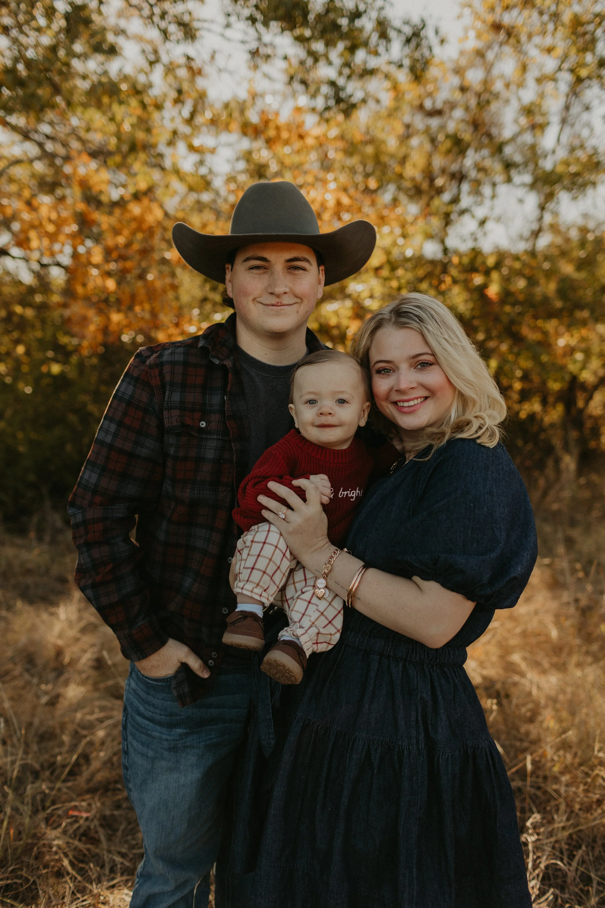 A smiling family of three, including a man wearing a cowboy hat, a woman, and a young child, outdoors during fall with autumn trees in the background.