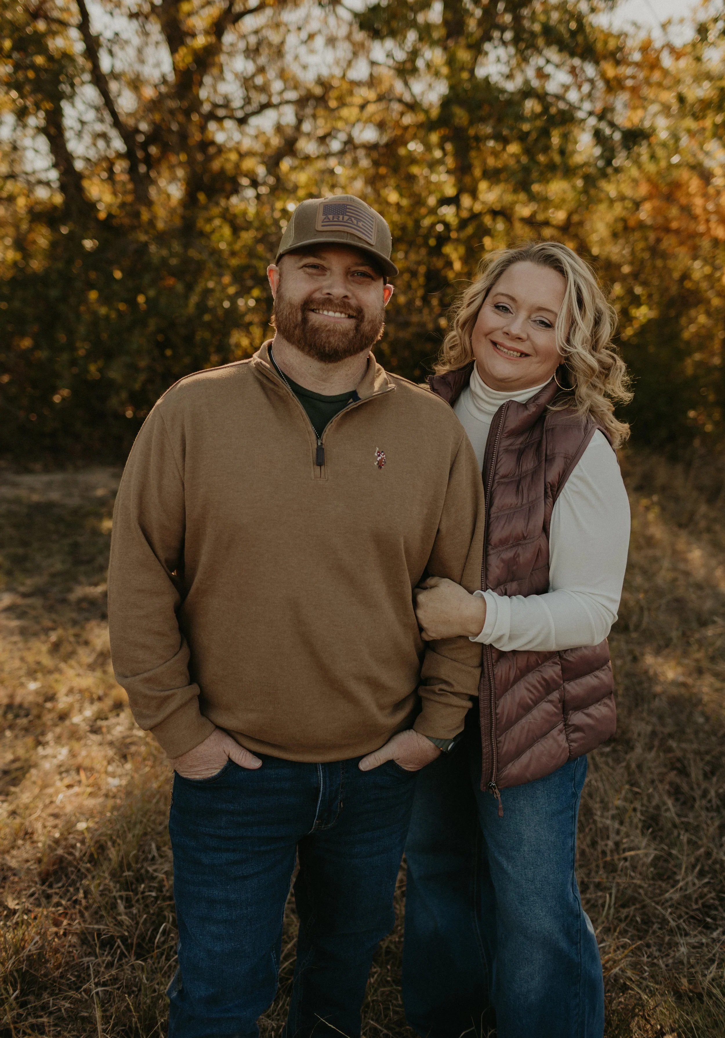 A happy couple standing outdoors in autumn, smiling for the camera with trees and golden leaves in the background.