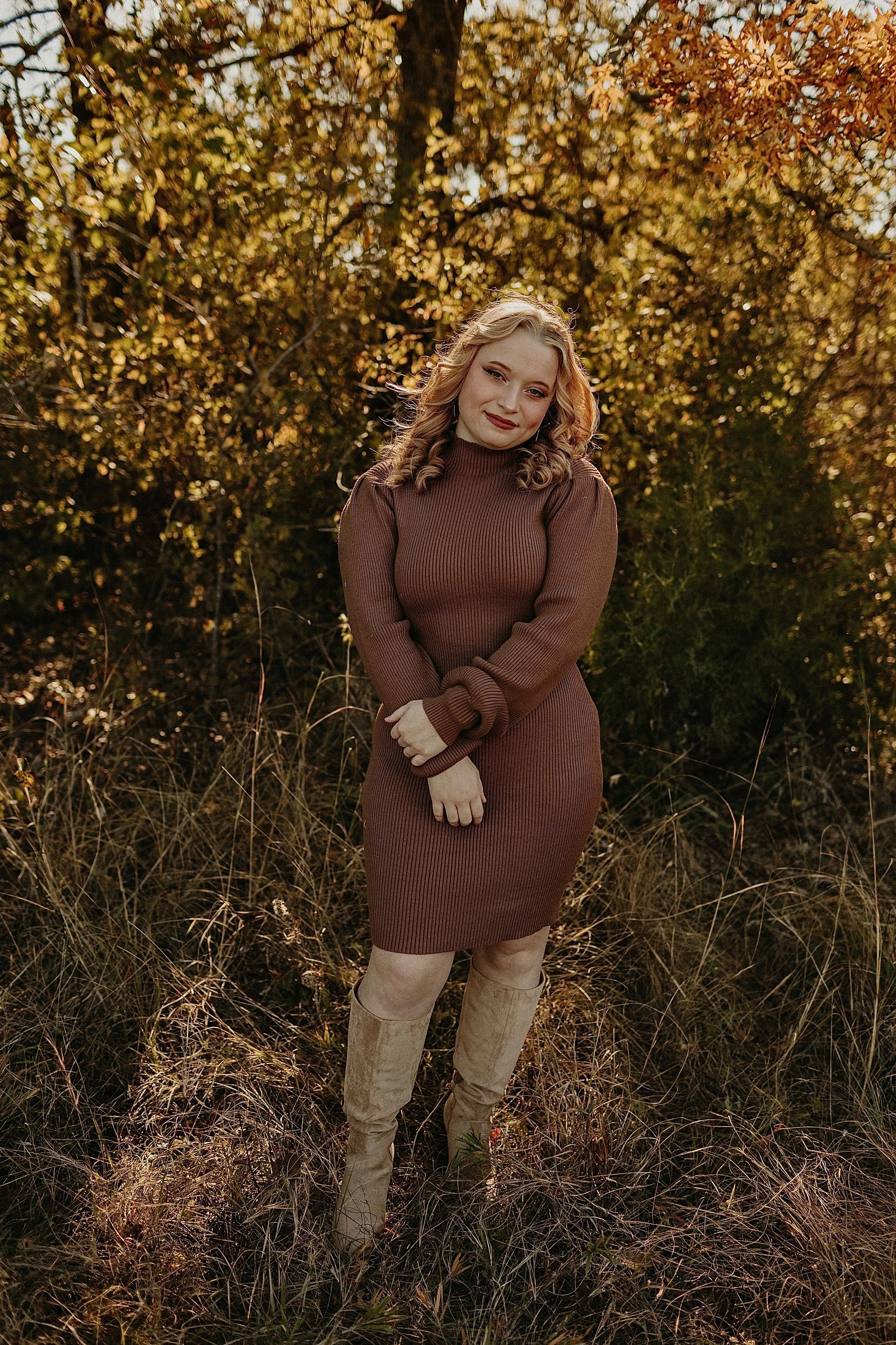 A young woman with blonde curly hair, wearing a brown ribbed sweater dress and beige boots, stands in a field of dry grass with autumn-colored trees in the background.