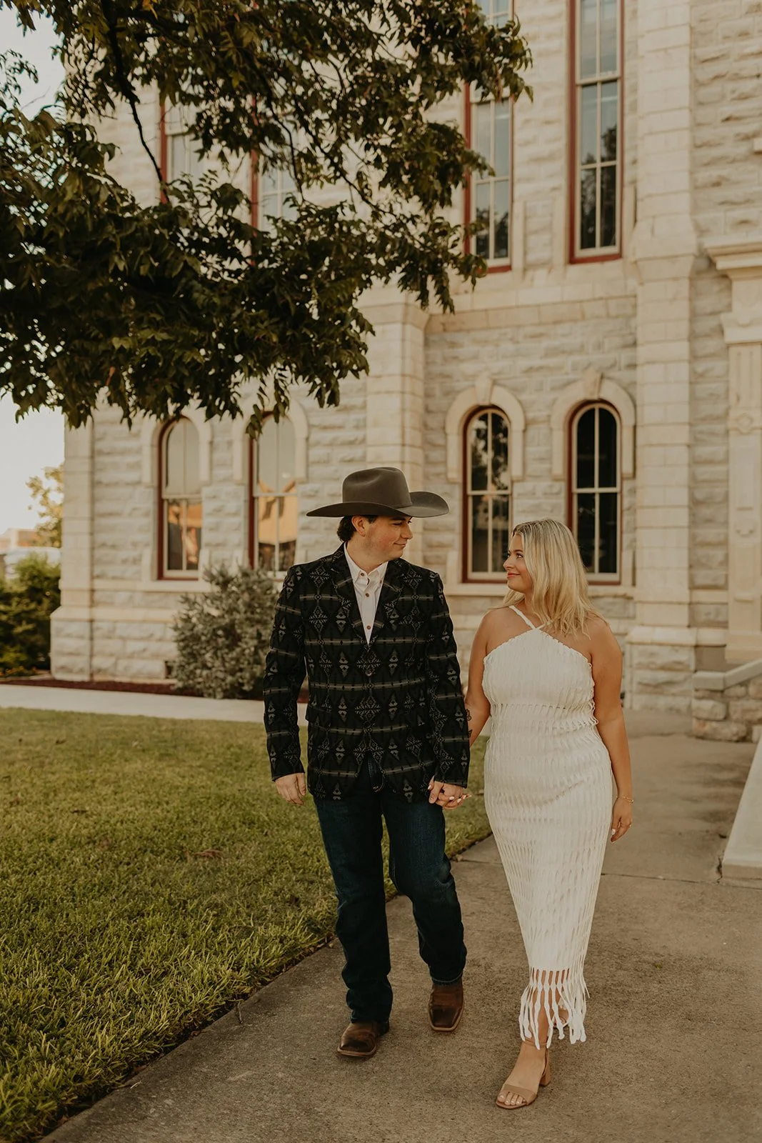 A man and woman walking hand in hand outside a stone building with arched windows, the man wearing a cowboy hat and patterned blazer and the woman in a white dress.