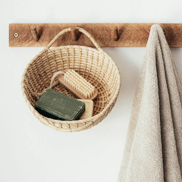 Organise & Harmonise. A woven basket hanging on a wooden wall hook containing a green bar of soap, a light-colored soap bar, and a scrub brush. A towel is hanging on the right side.