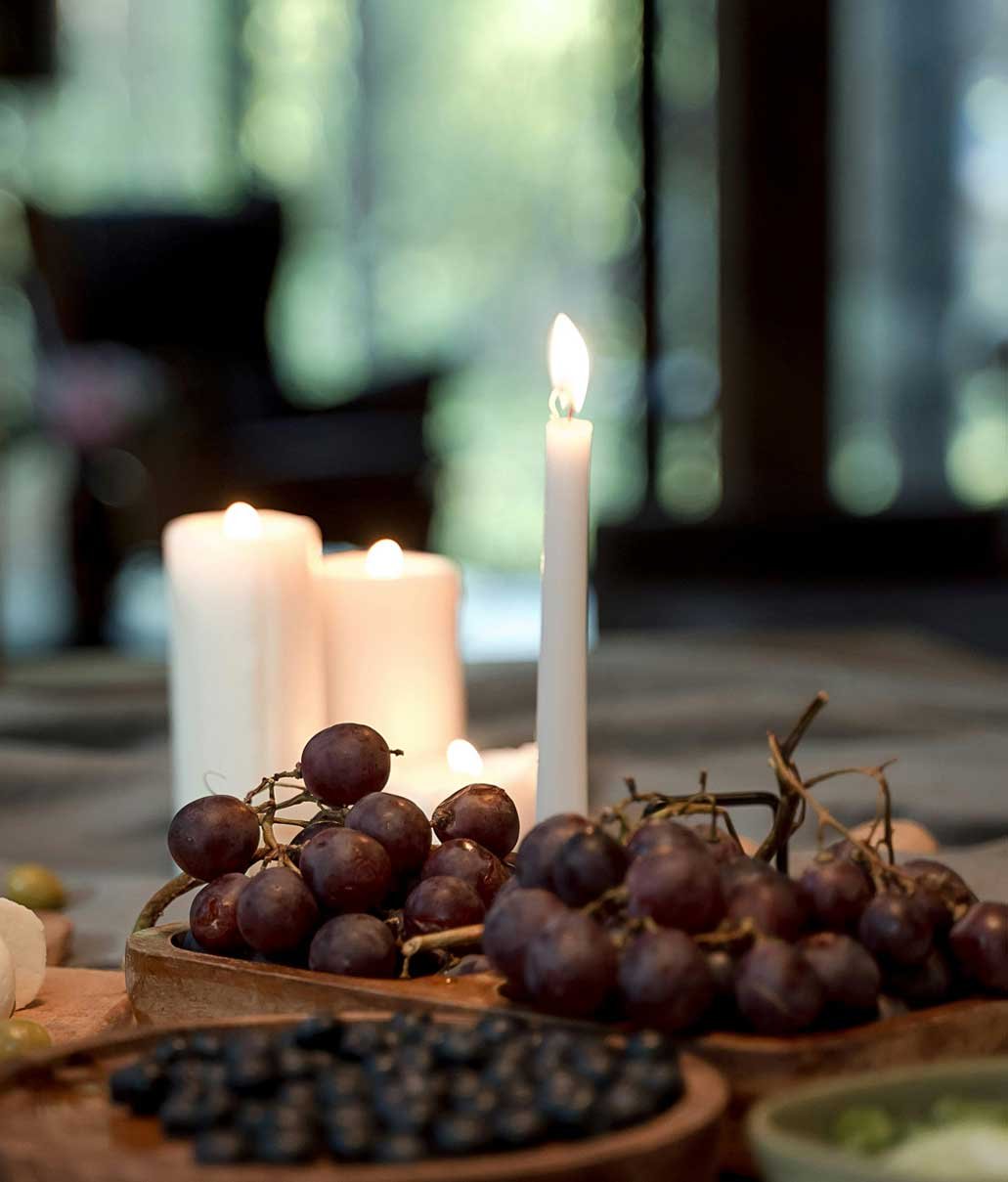 Close-up of a table with red grapes, candles, and a green dish, with blurred candles and windows in the background.