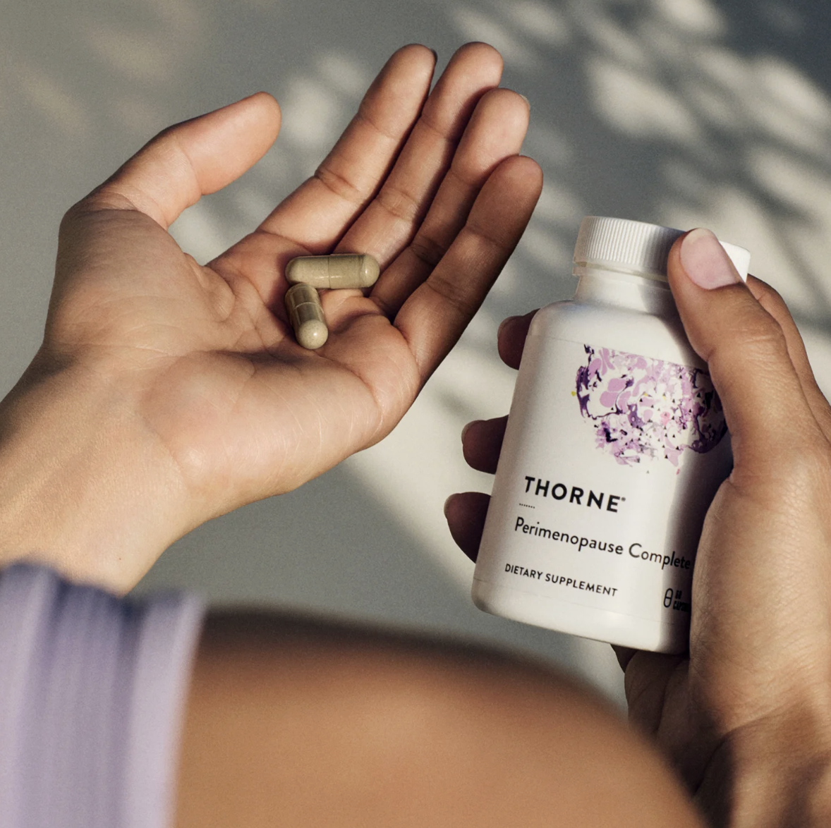 A person holding a white bottle labeled 'THORNE Perimenopause Complete Dietary Supplement' in one hand and three beige capsules in their other hand.