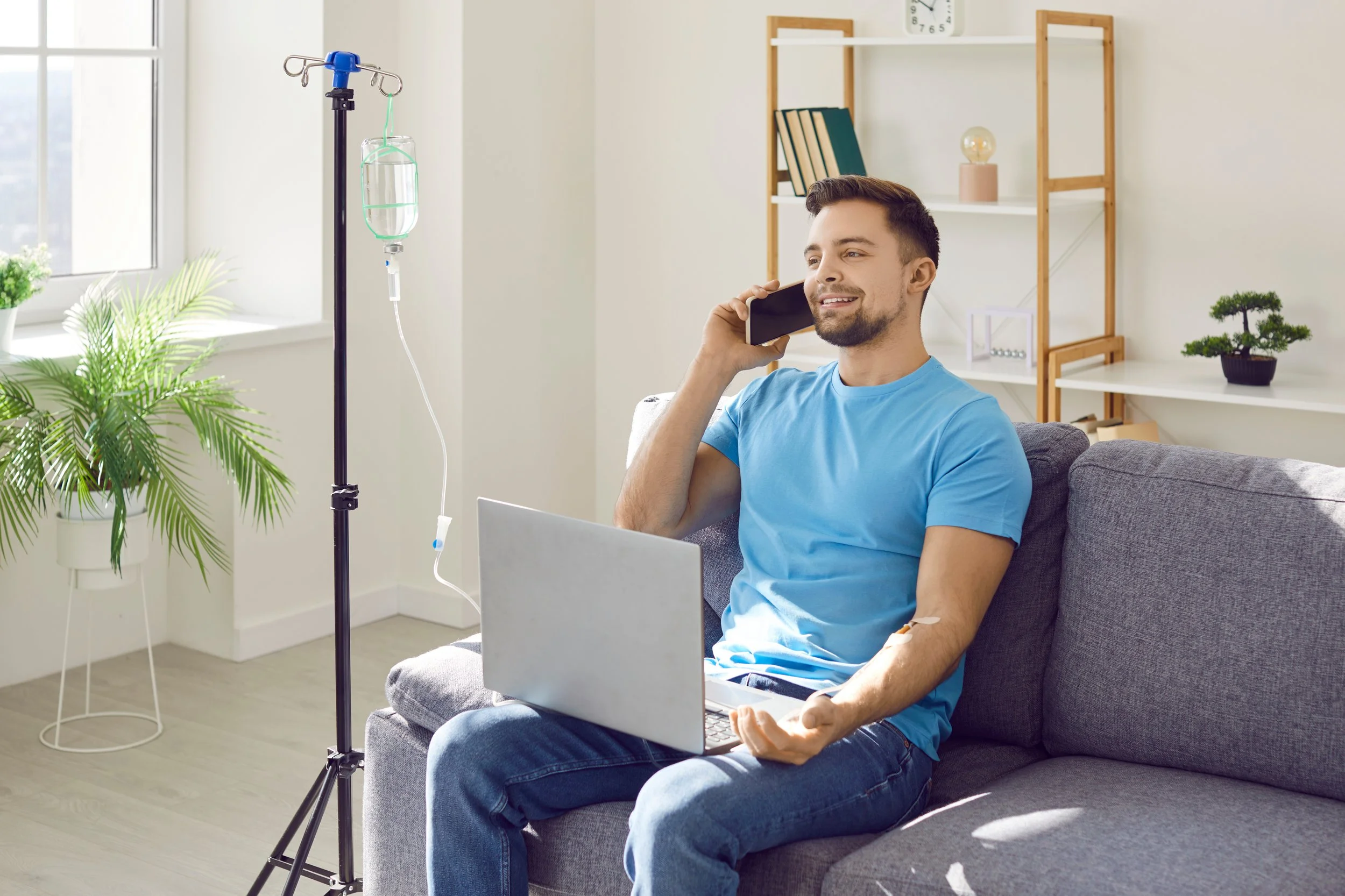 A man in a blue shirt sitting on a couch, talking on a cell phone, with a laptop on his lap and an IV drip stand nearby in a well-lit room with plants and shelves.