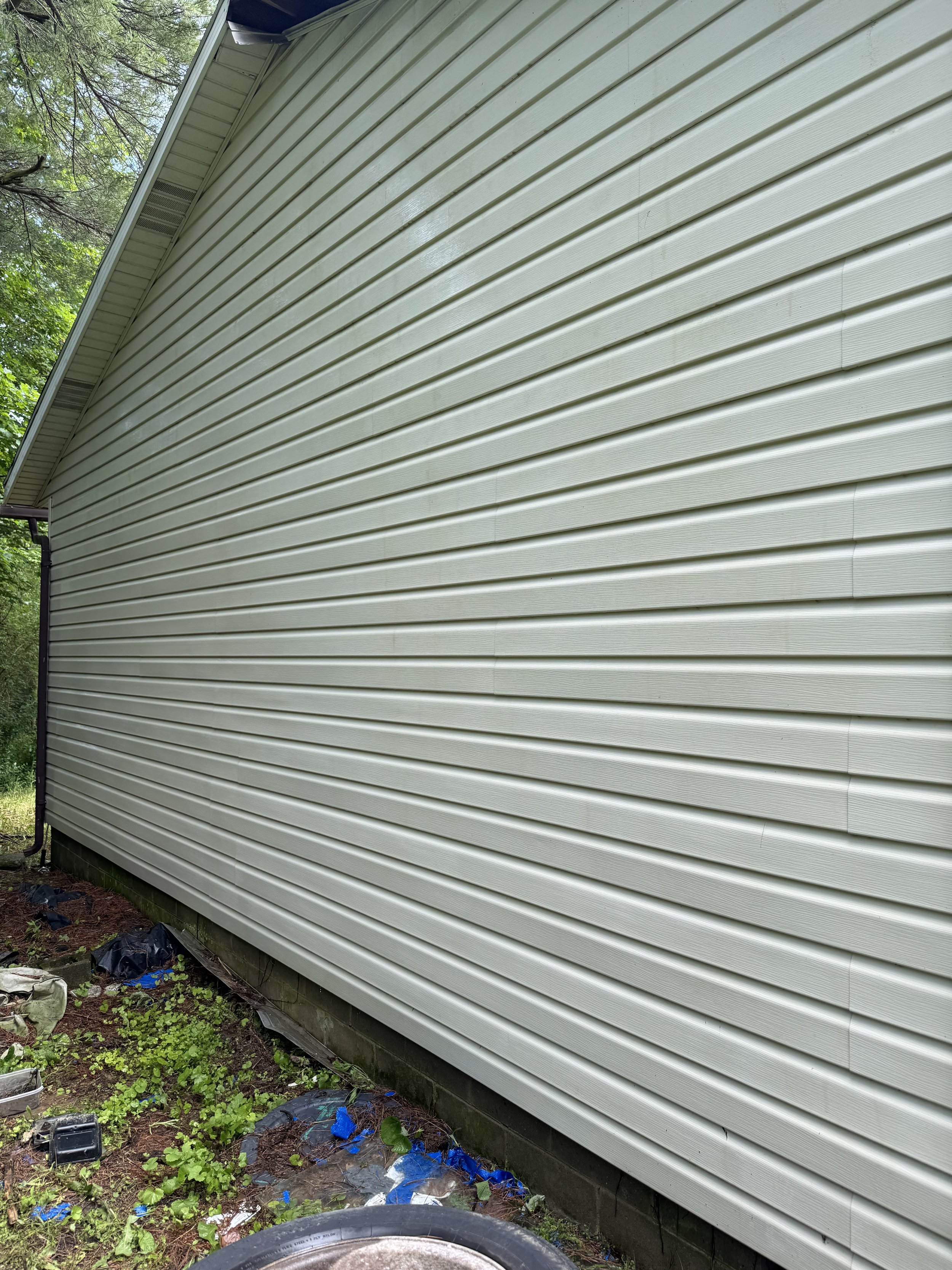 Side of a house with beige siding and some debris, trash, and green plants on the ground near the base of the house.