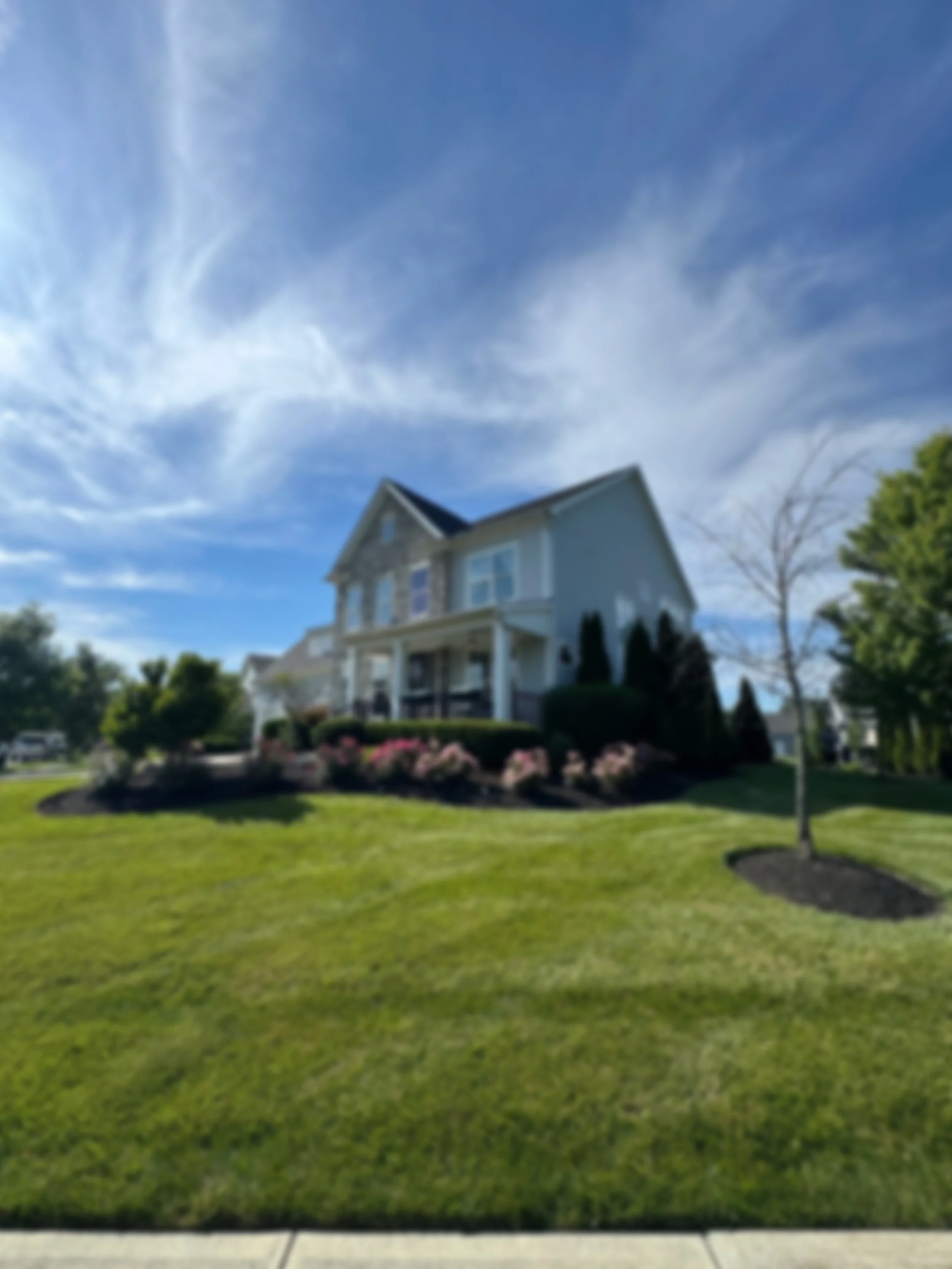 A two-story house with a front porch, surrounded by a well-maintained lawn and bushes, under a partly cloudy blue sky. A tree with a circular mulch bed is in the foreground on the right.