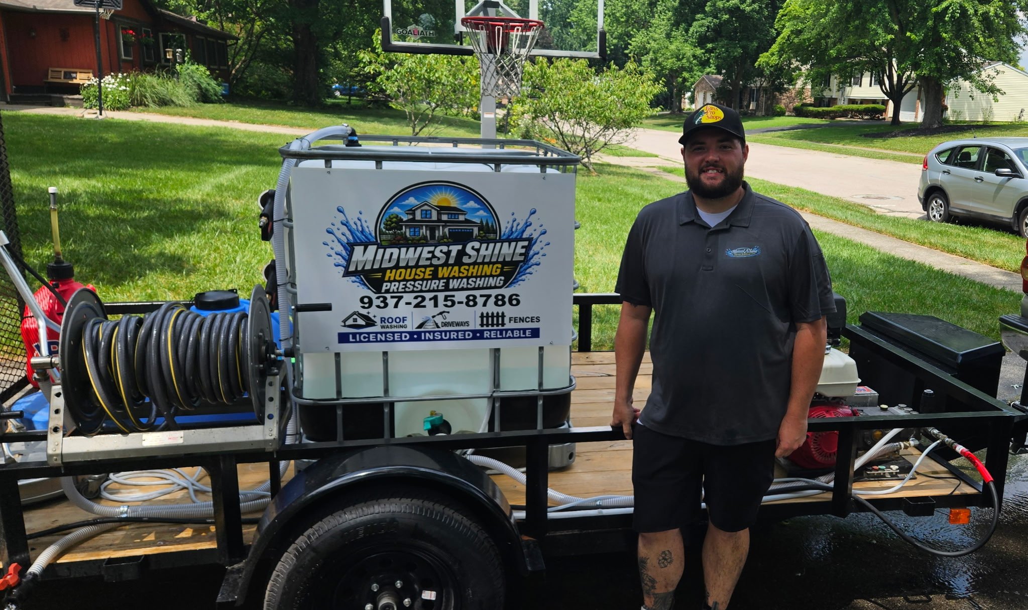 A man wearing a black polo shirt and a black cap with a yellow logo stands next to a house washing pressure machine on a wooden platform in a suburban area with houses and green trees in the background. The pressure washing equipment includes hoses and a water reel, with a sign displaying the business name 'Midwest Shine House Washing Pressure Washing' along with a phone number and services offered like roof washing, driveway cleaning, and fencing.