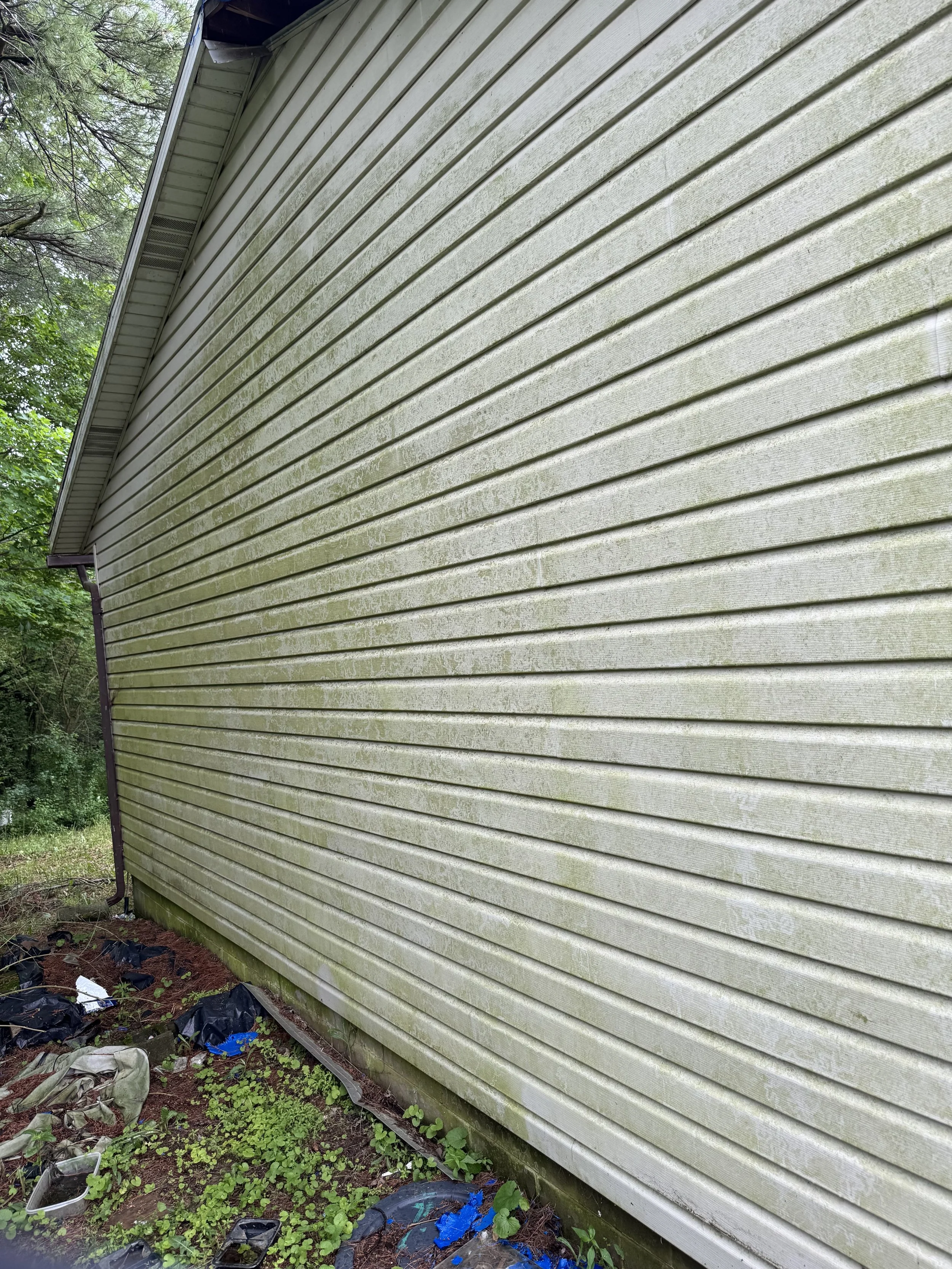 The side of a house with dirty, weathered beige vinyl siding and some scattered trash on the ground.