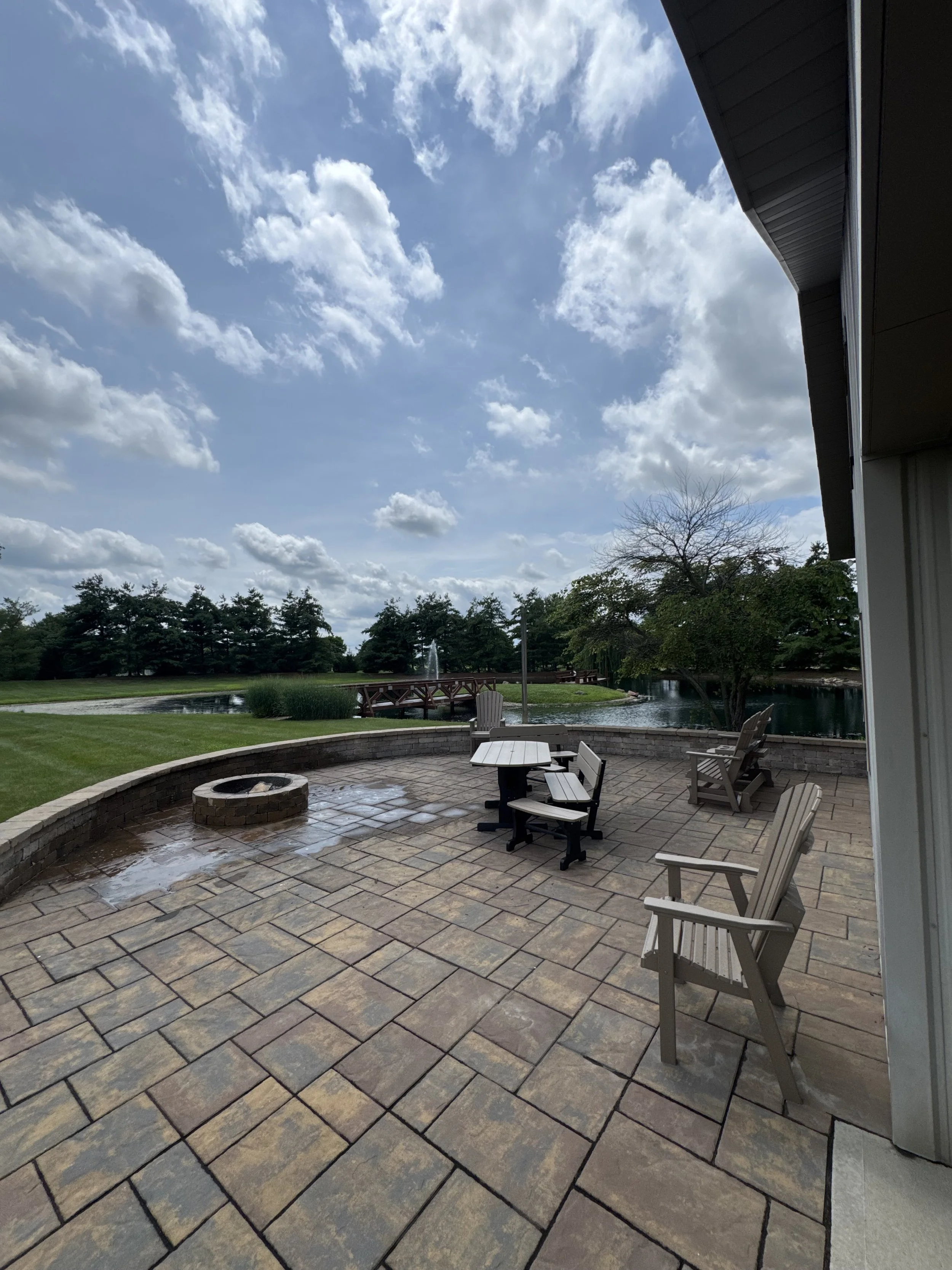 A backyard patio with a fire pit, outdoor chairs, a table, a pond with a fountain, and a lawn with trees under a partly cloudy sky.
