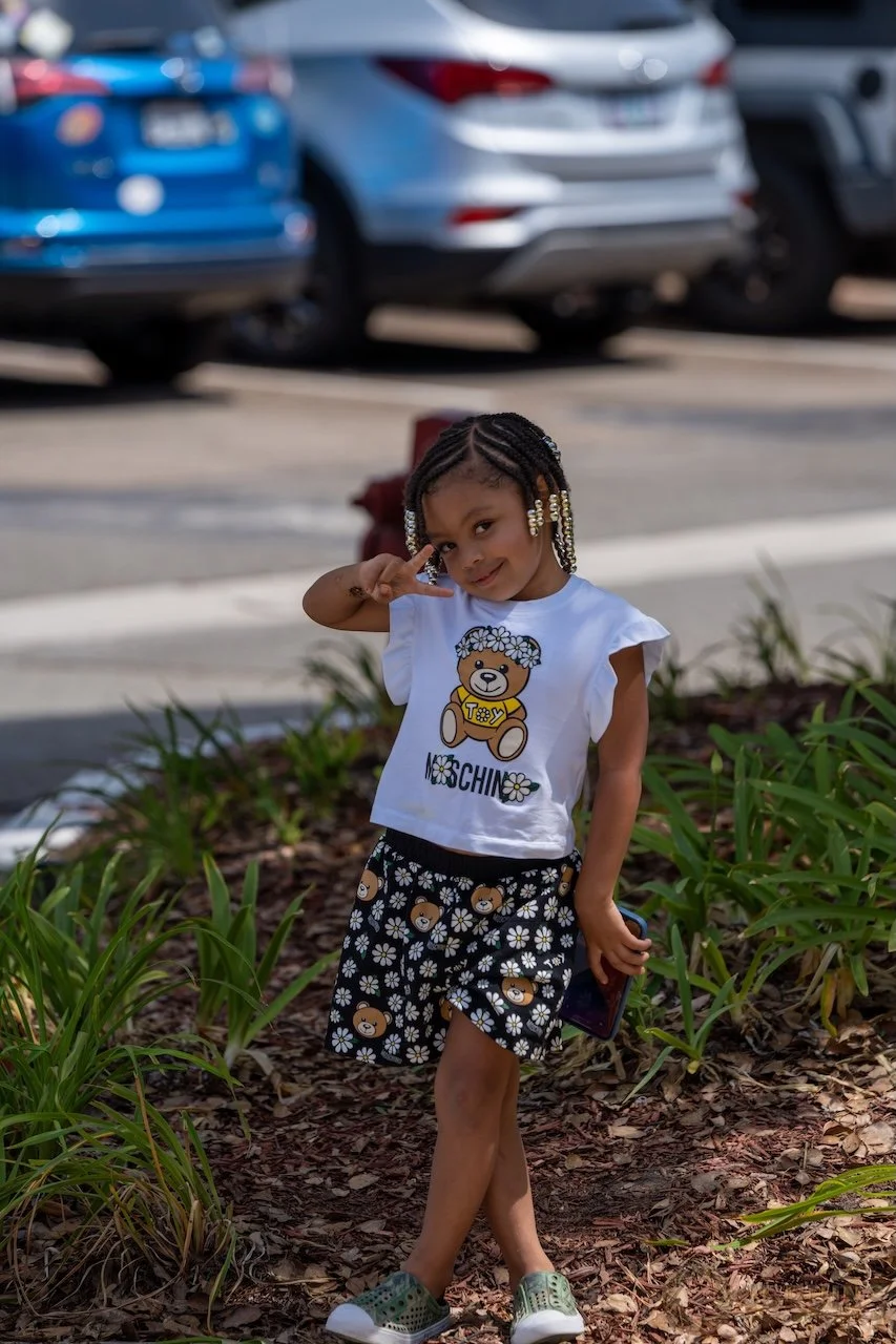 A young girl with braided hair, wearing a white T-shirt with a teddy bear design and the word 'MOSCHINO,' floral shorts with teddy bear patterns, and green slip-on shoes, standing outdoors among green plants, holding a phone, and making a peace sign with her right hand.