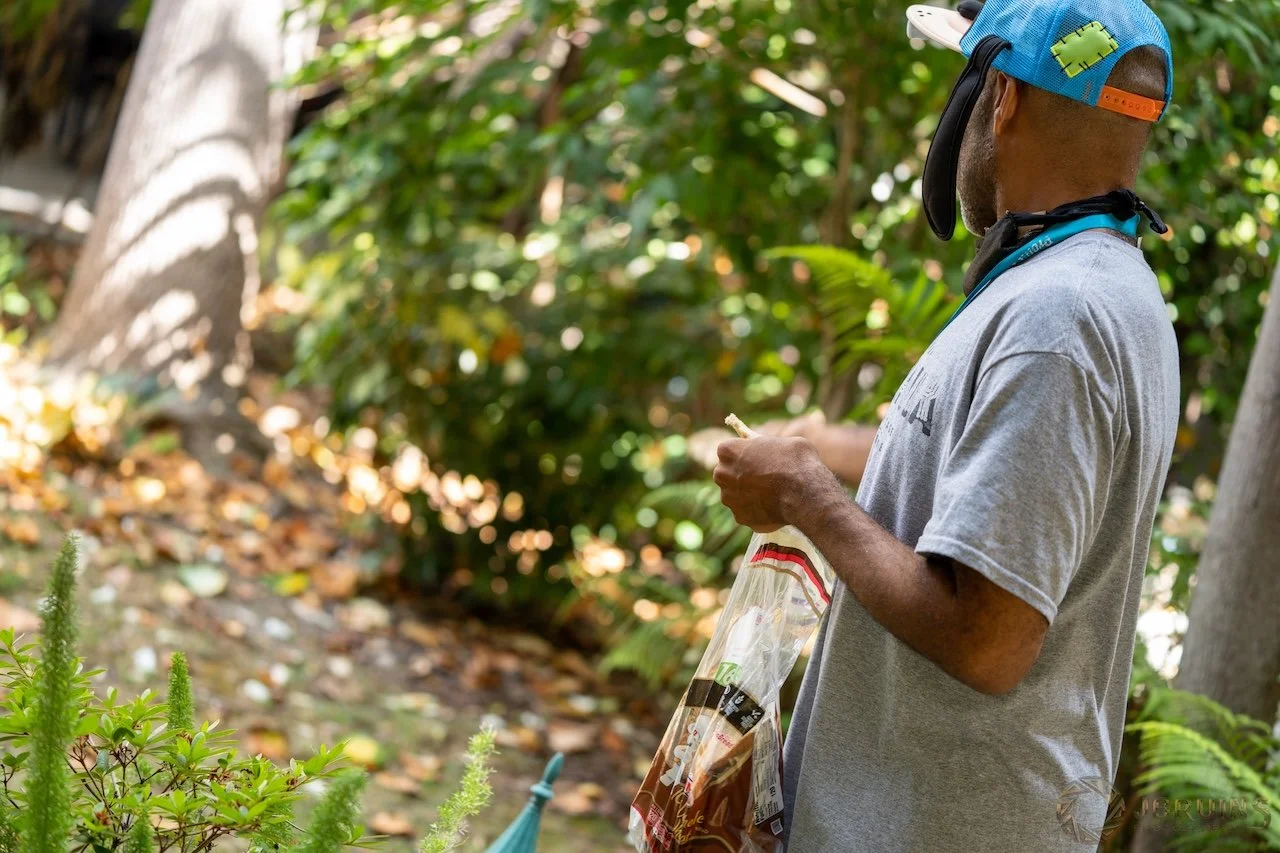 A man wearing a gray shirt, a colorful baseball cap with a puzzle piece patch, and a face mask hanging around his neck, standing outdoors in a wooded area, holding a plastic bag of snacks.