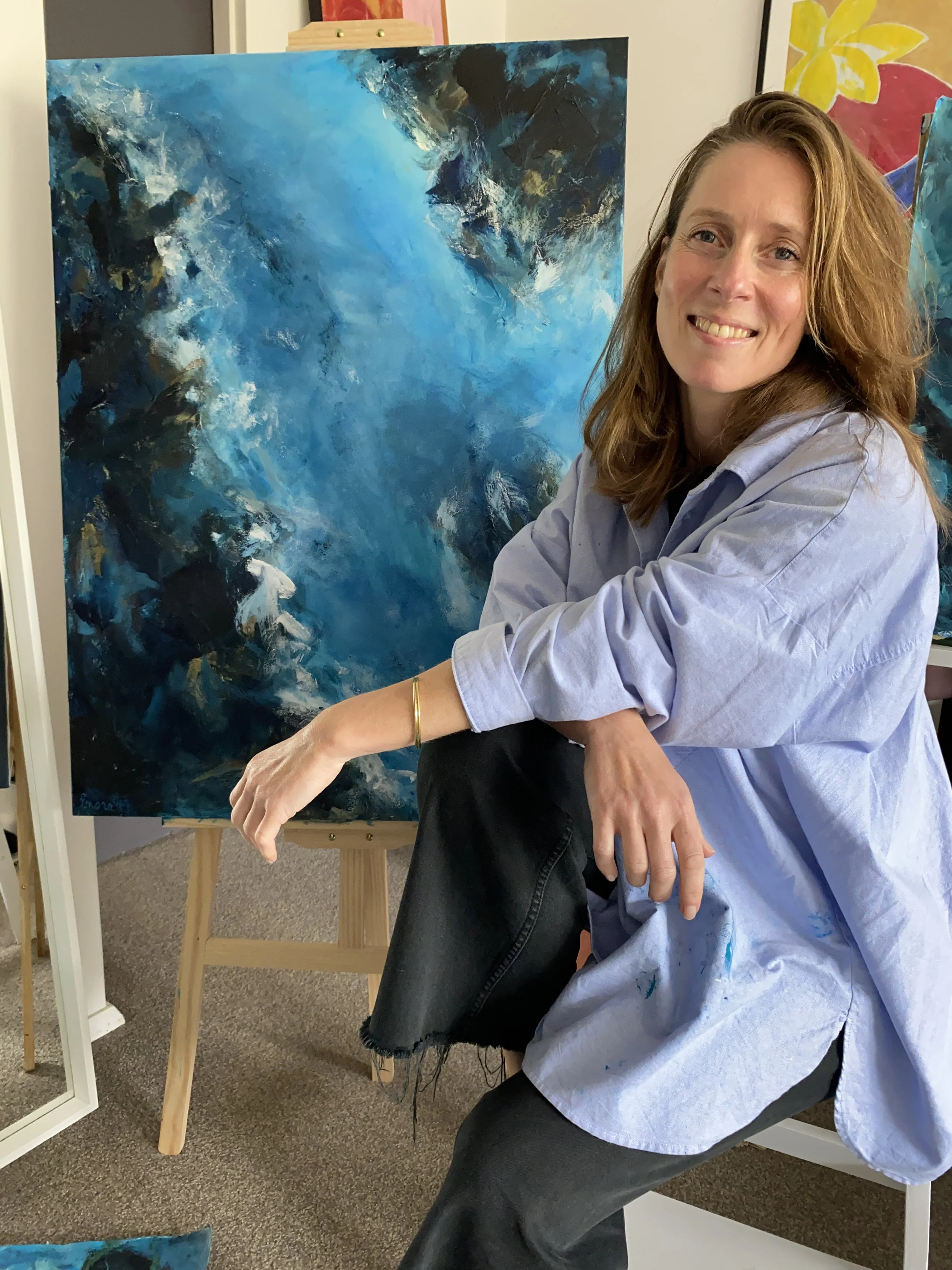 A smiling woman with wavy red hair sitting in front of an abstract blue painting on an easel. The woman is wearing a light blue shirt and dark pants, and she appears to be in an art studio or gallery.