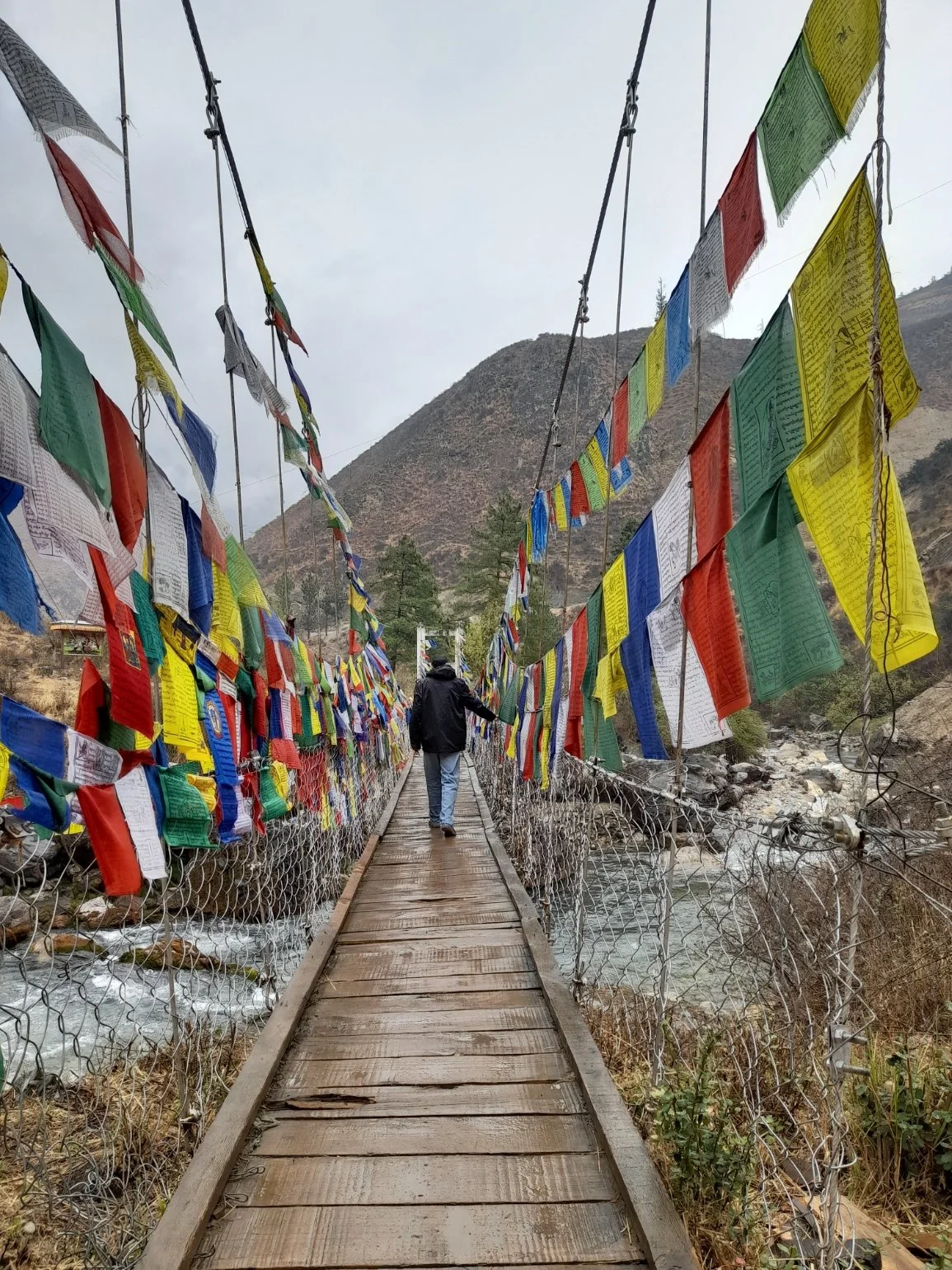 A person walking across a suspension bridge decorated with colorful prayer flags with a mountain and trees in the background.