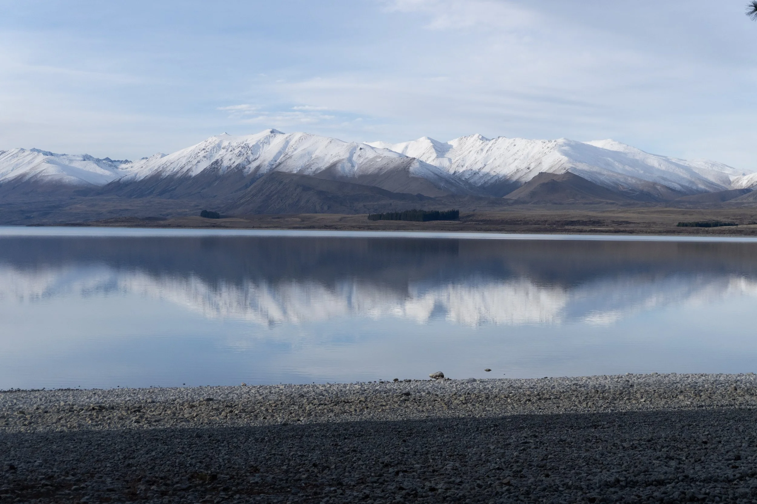Snow-capped mountains reflected in a calm lake with a rocky shoreline in the foreground.