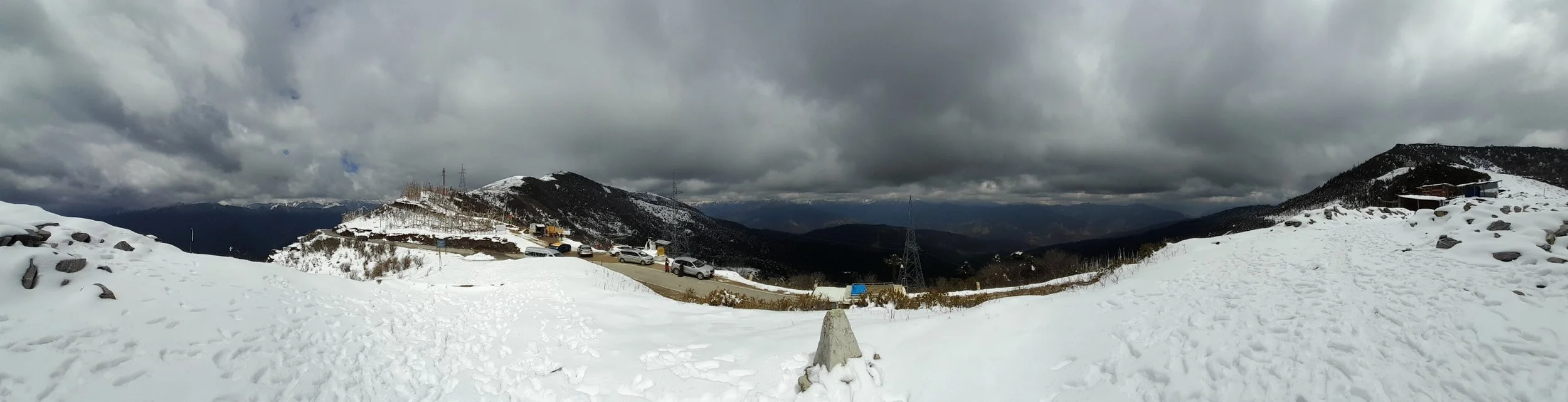 Snow-covered mountain landscape with parked cars, a winding road, and cloudy sky.