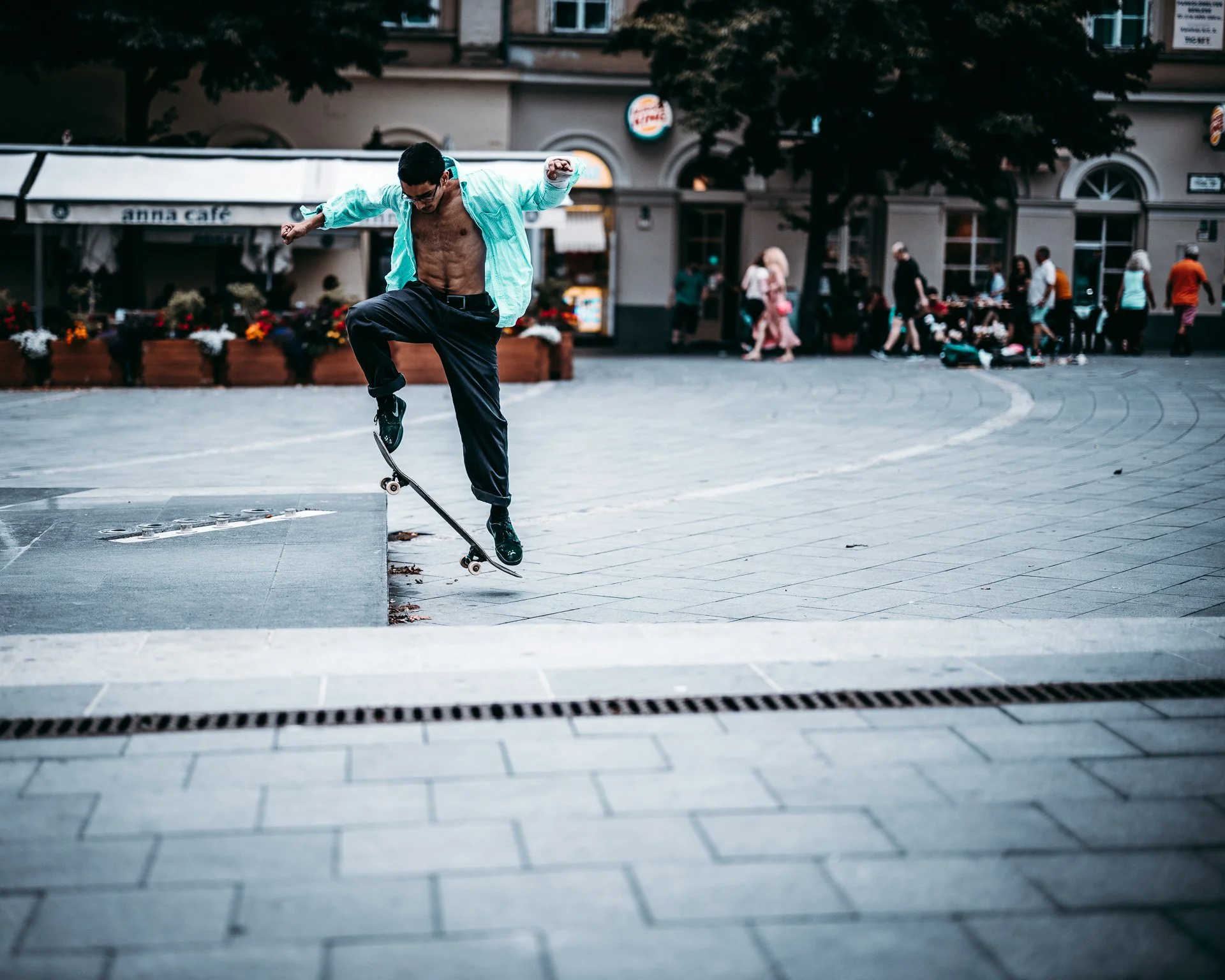 A shirtless man with glasses skateboarding on an urban street, wearing black pants and black shoes, with a crowd of people and storefronts in the background.