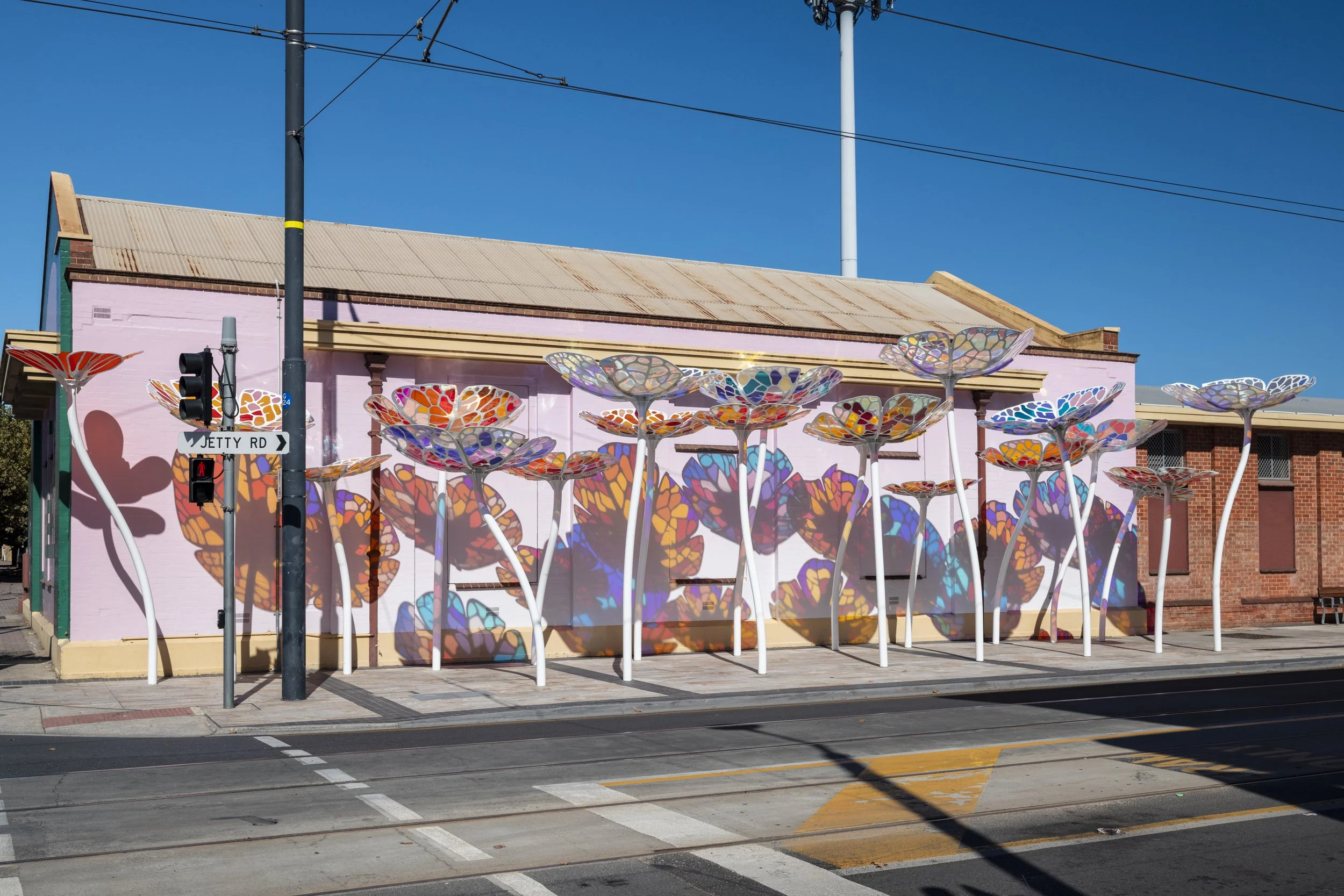 Colorful butterfly-shaped art installation on the side of a pink building with a rusty roof, blue sky, and street with a traffic light and street signs nearby.
