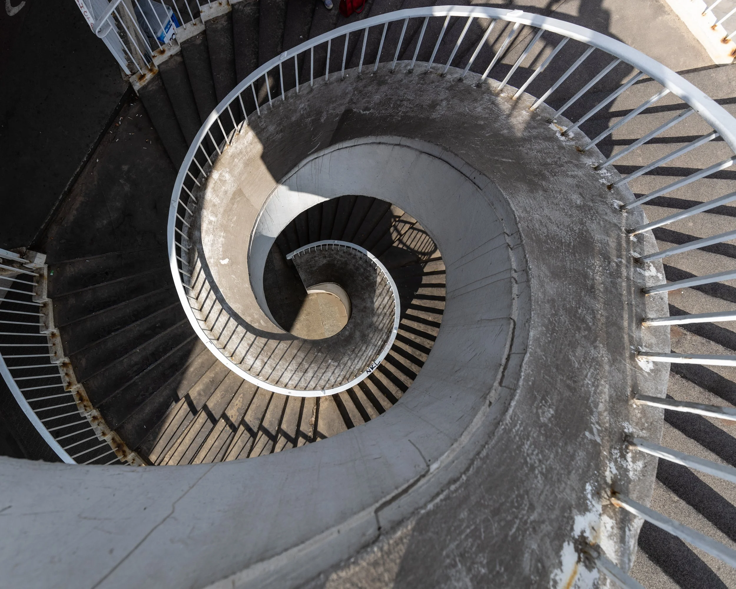 Spiral staircase viewed from above, with white railings and dark steps, showing rust and weathering.