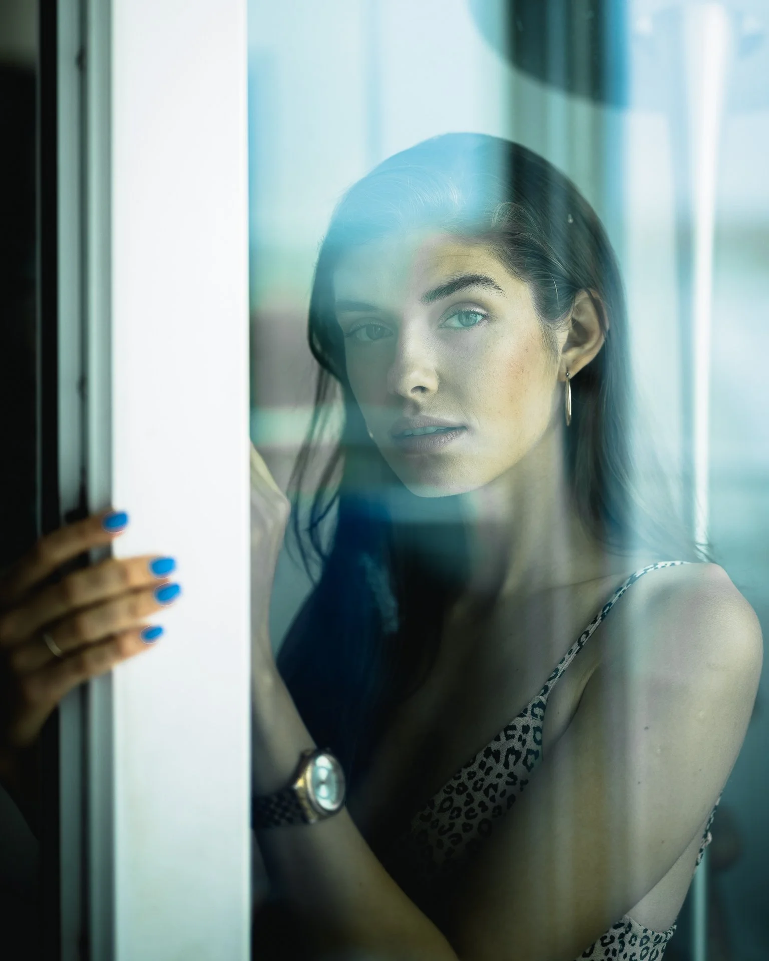 A woman with blue eyes and dark hair, wearing a watch and earrings, peering through a glass window or door with a blurred background.