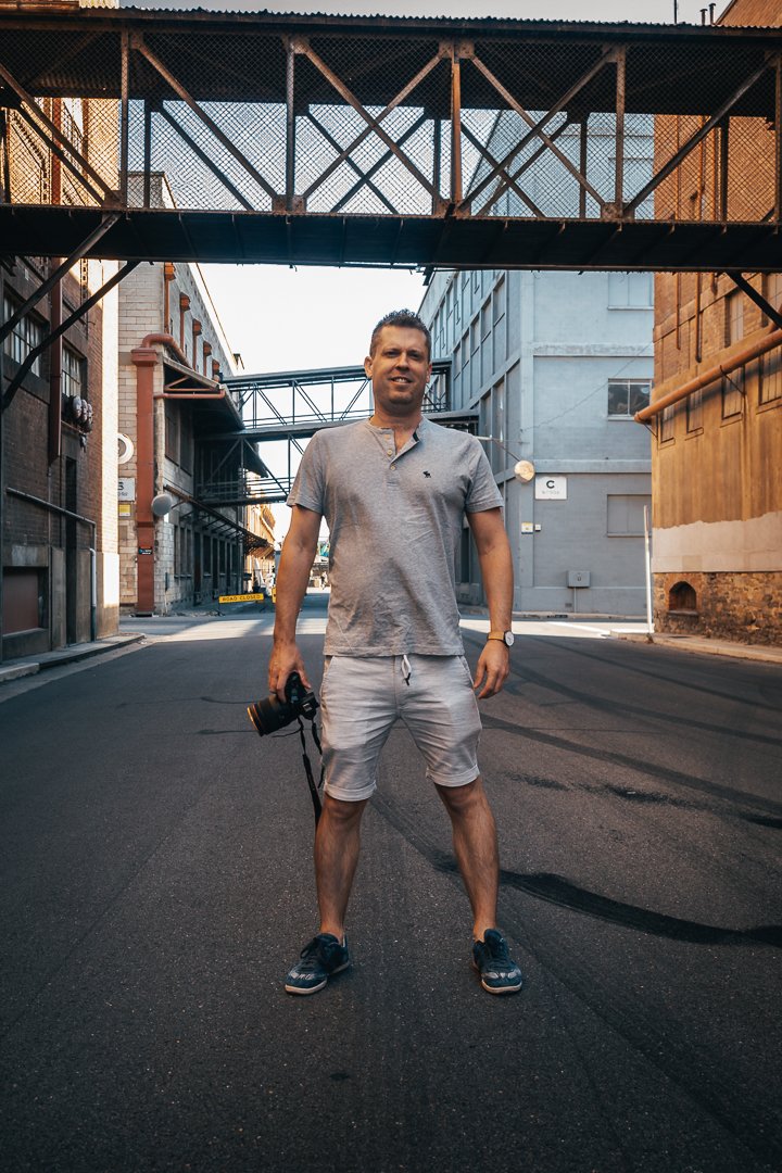 Ben Owens standing in the middle of a city street holding a camera, wearing a gray t-shirt, shorts, and sneakers, with industrial buildings and sky bridges in the background.