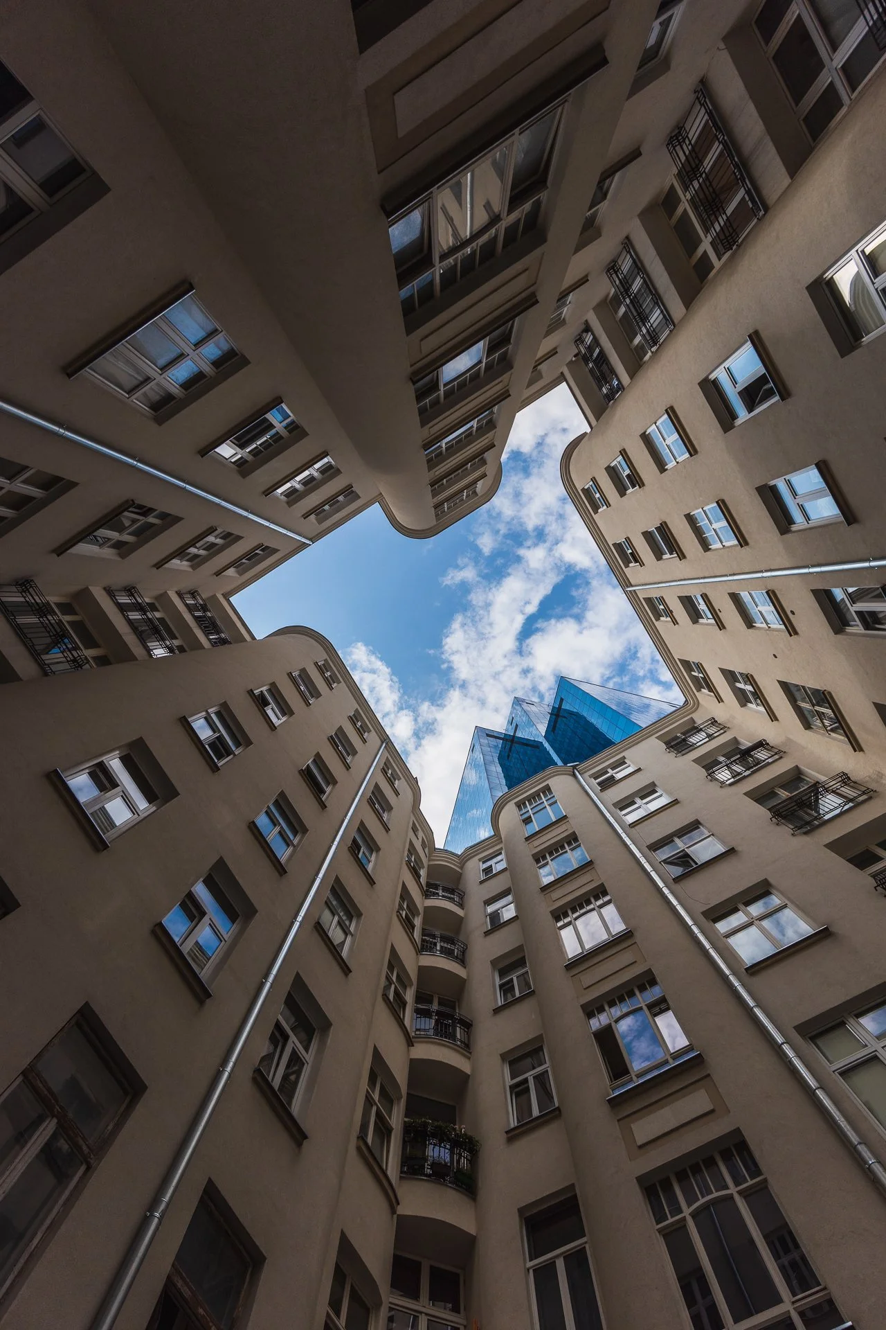 Looking up from the courtyard of a multi-story residential building, showing beige walls, numerous windows, black metal balconies, and a blue glass skyscraper in the background against a partly cloudy sky.