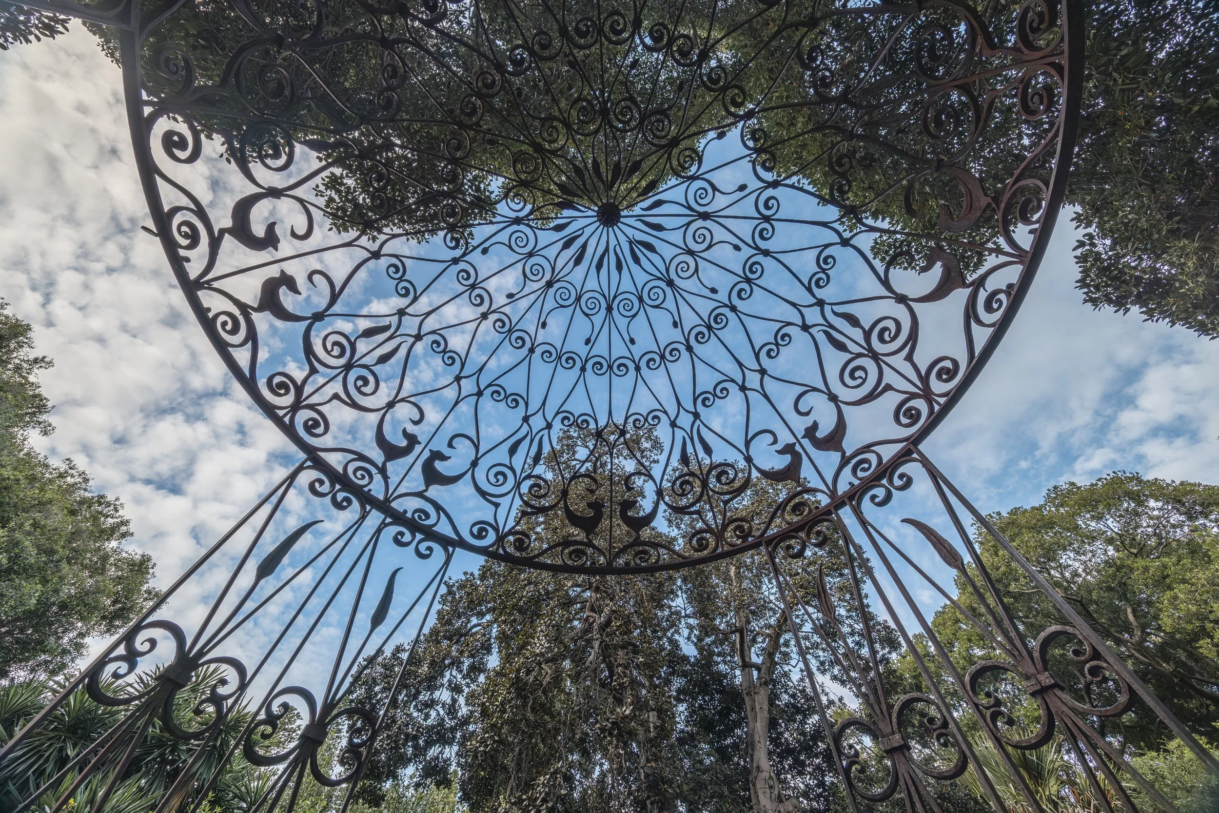 Ornate wrought iron gate with intricate scrollwork design, viewed from below, with trees and a partly cloudy sky visible in the background.