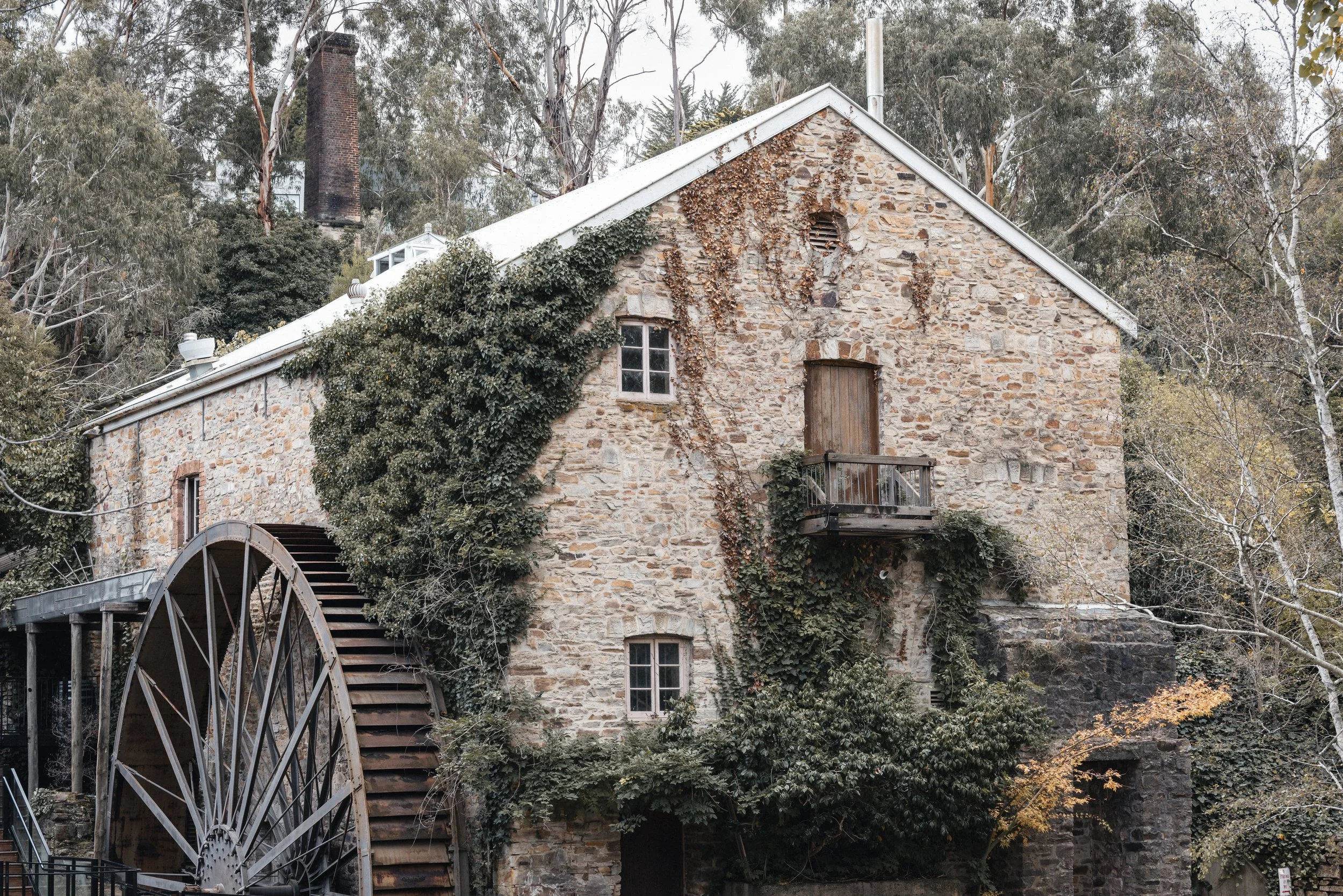 A rustic stone building surrounded by trees with some ivy growing on the walls and a water wheel at the front.