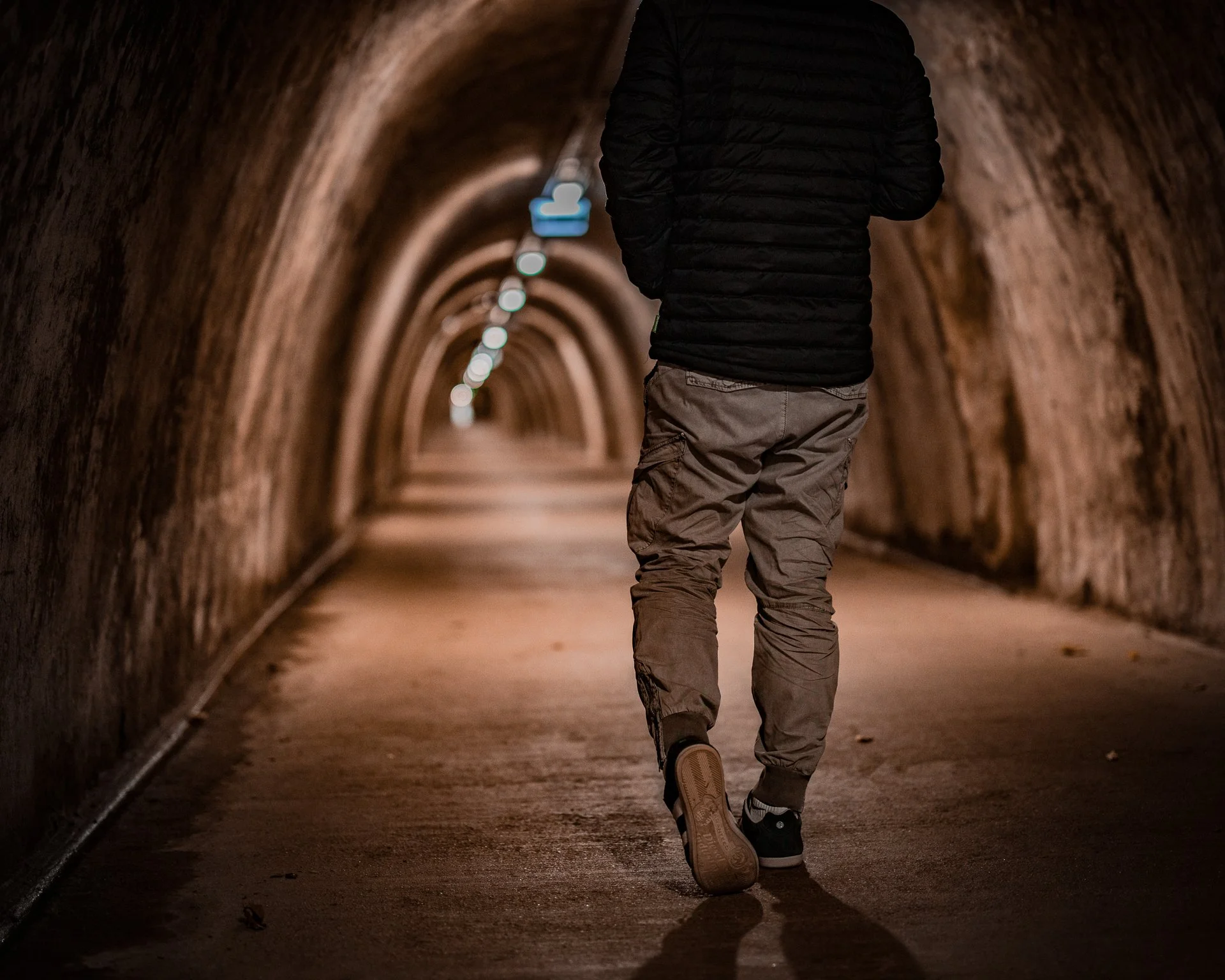 A person walking away down a tunnel with arched ceiling, wearing a black jacket and gray cargo pants, on a dirt or gravel surface.
