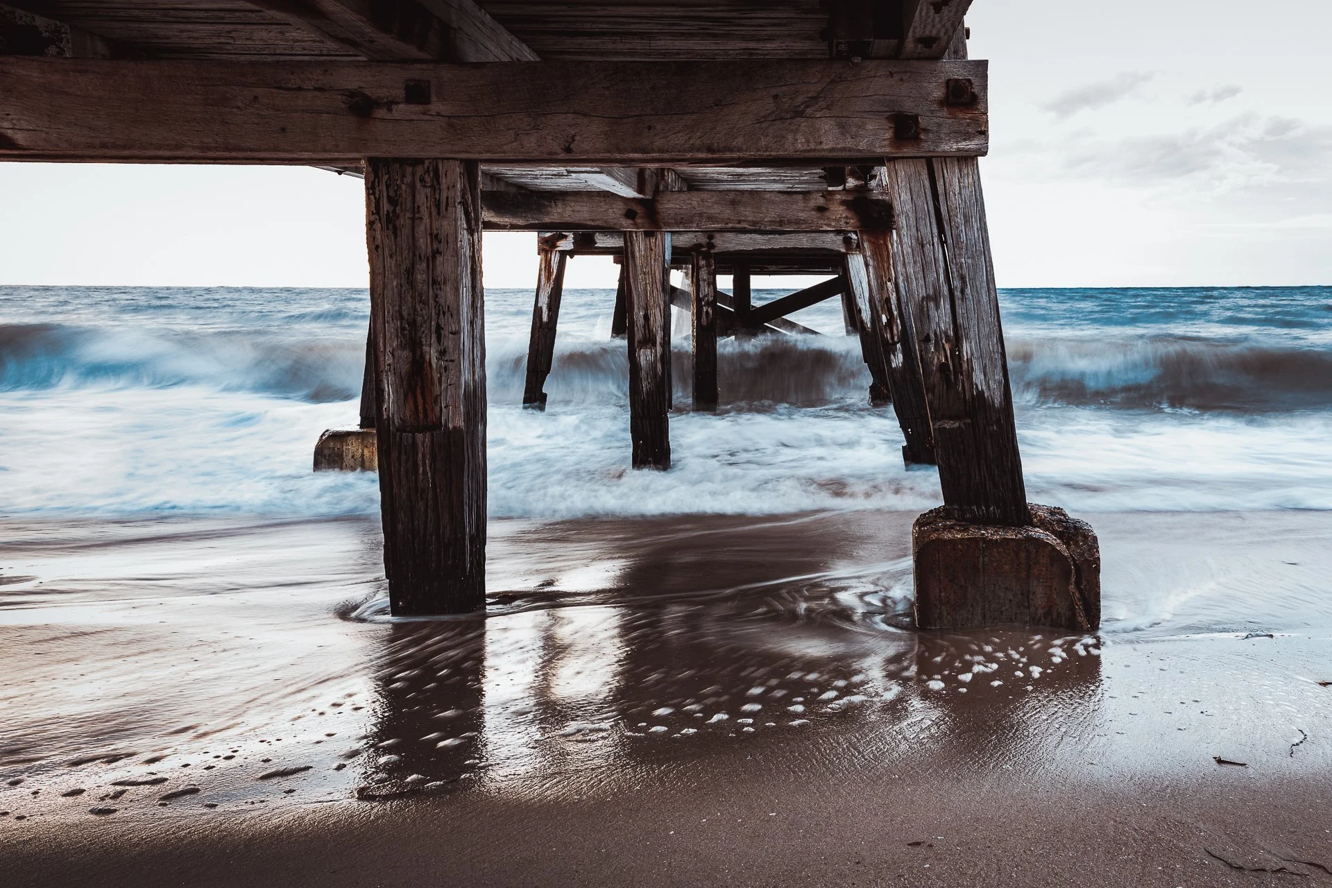 View underneath a wooden pier at the beach with waves crashing against the pilings and wet sand in the foreground.