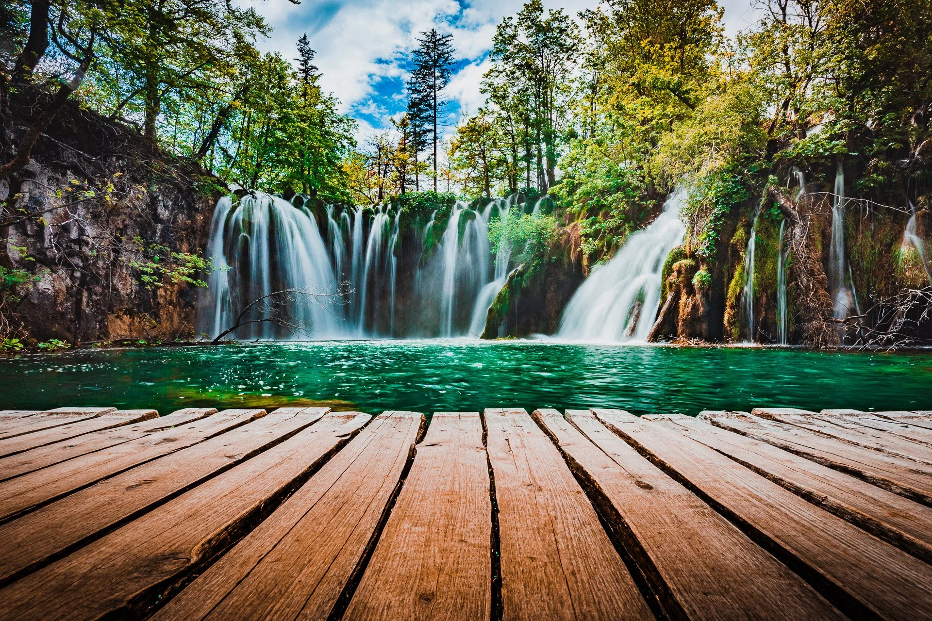 A scenic view of a waterfall flowing into a clear green pond surrounded by lush green trees under a partly cloudy sky, with a wooden dock in the foreground.