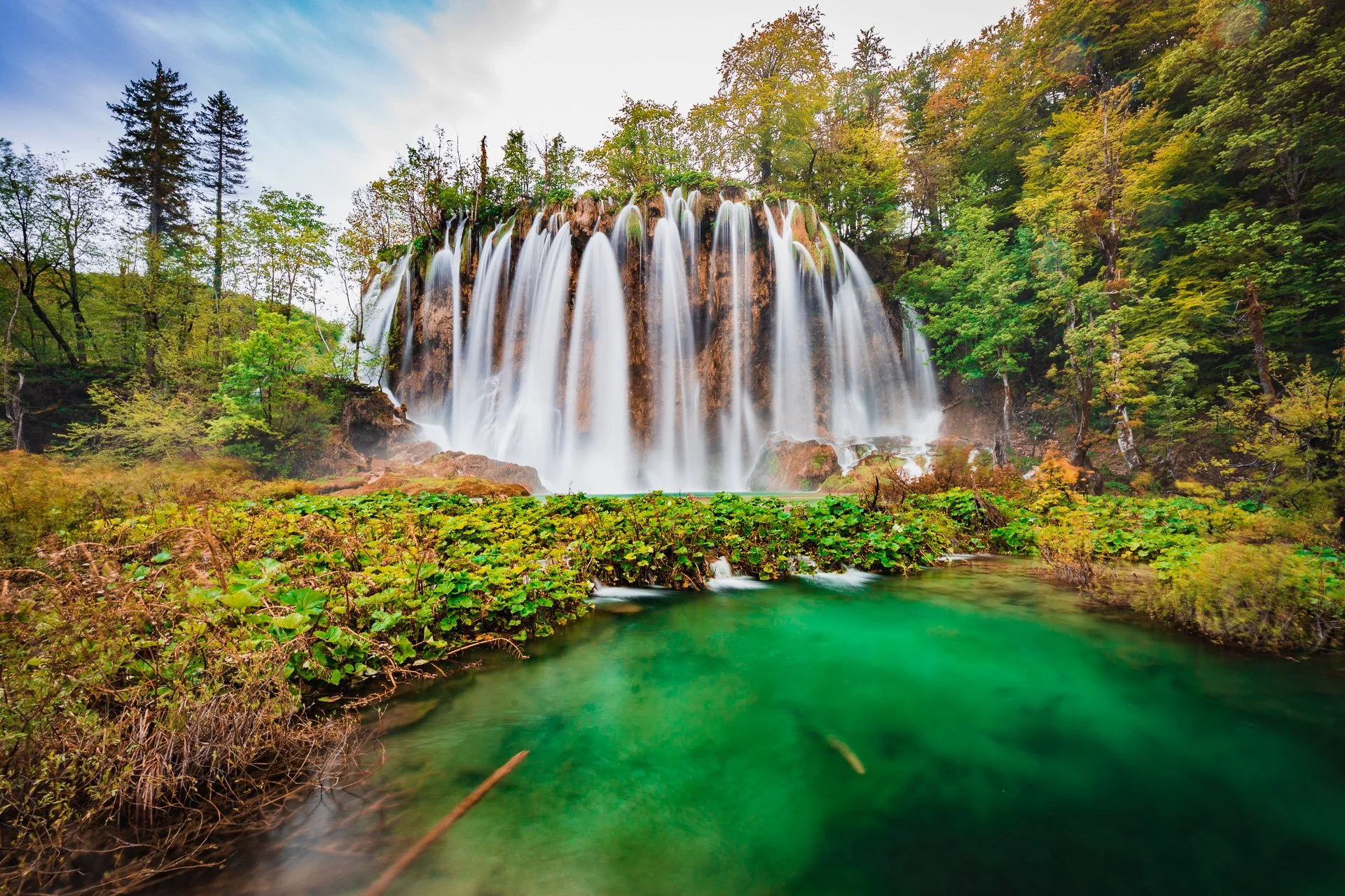A waterfall flowing over rocks surrounded by lush green trees and foliage, with a calm green pool at the base and a partly cloudy sky above.