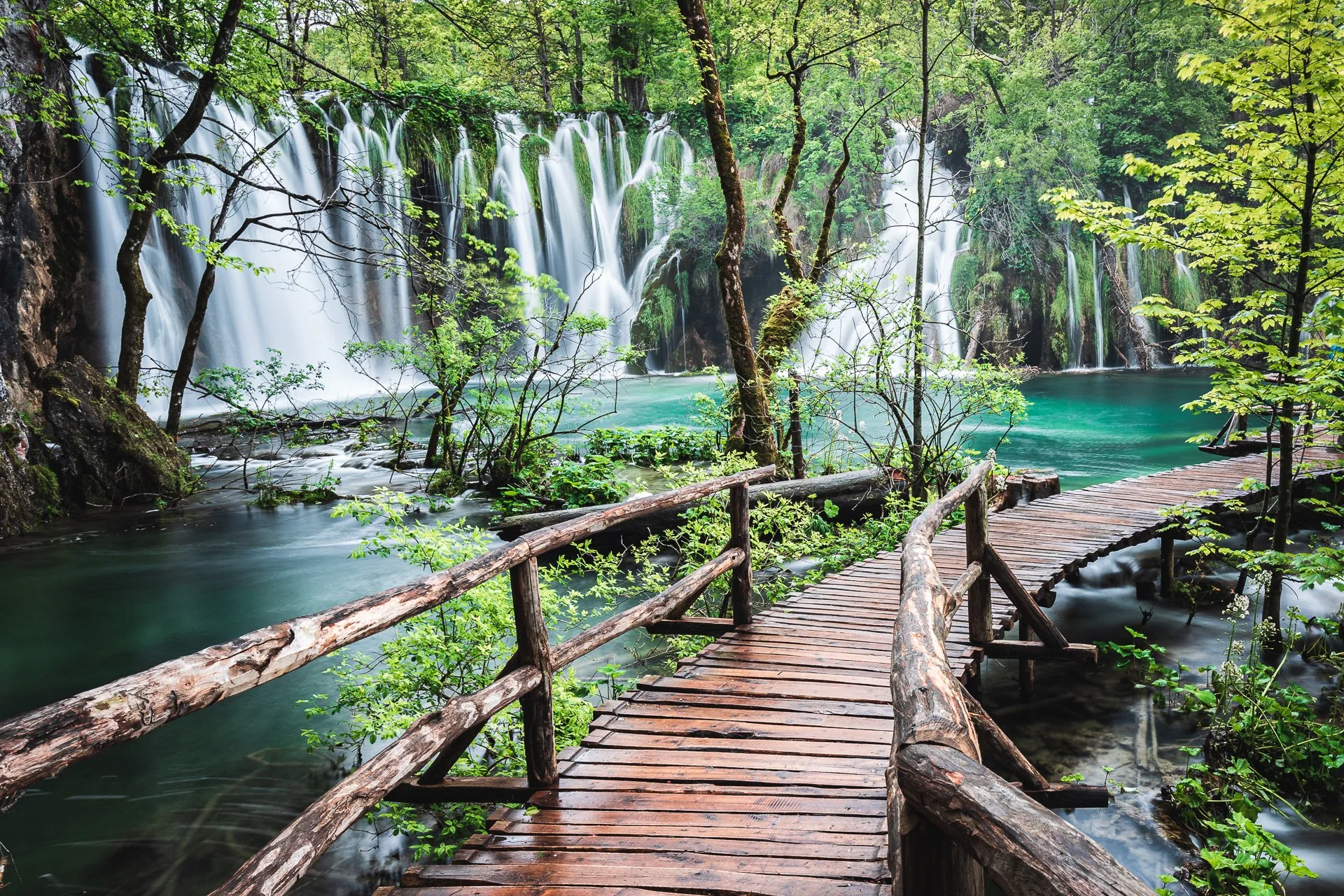 A scenic view of a forest with tall trees and a flowing waterfall cascading into a turquoise river. A wooden boardwalk with rustic railings runs along the water's edge.