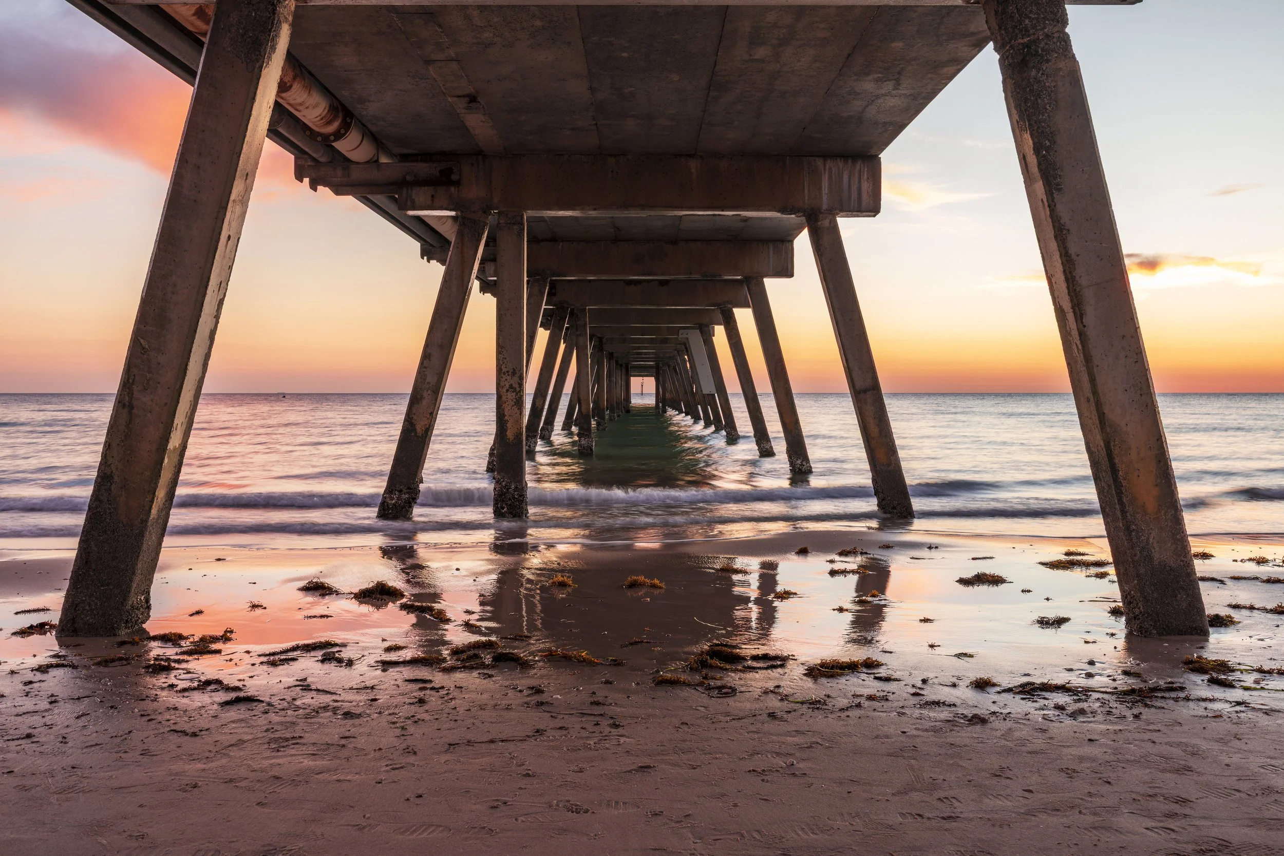 Underneath a pier at sunset with the sky showing pastel pink, orange, and purple hues over the ocean. The pier's structural supports are rusted and extend into the water, with waves gently lapping the shore, which is scattered with seaweed.