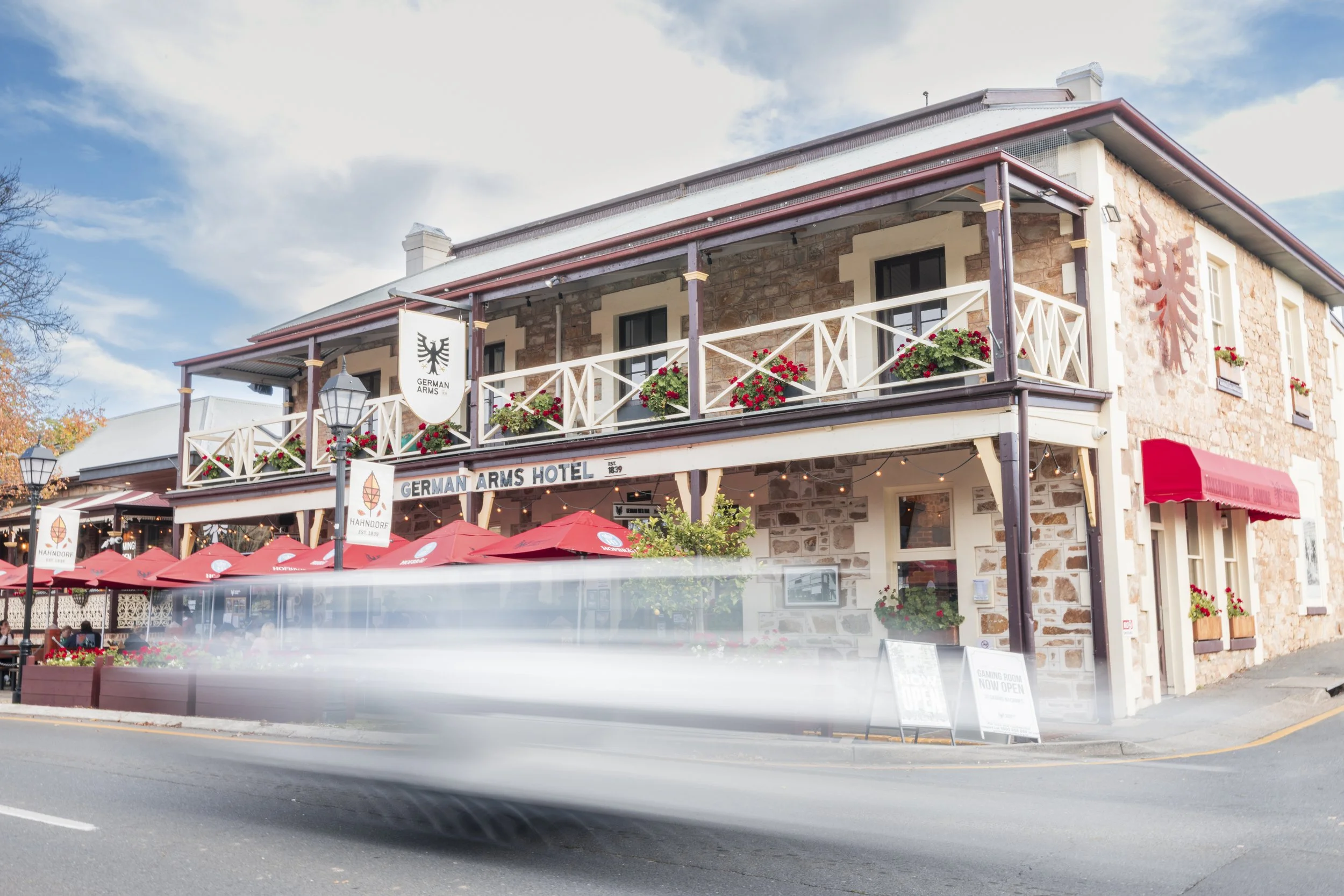 A two-story building with a sign that reads 'German Arms Hotel' on the front facade, decorated with red flowers on the balcony, under a partly cloudy blue sky. Outdoor seating with red umbrellas is visible at street level.