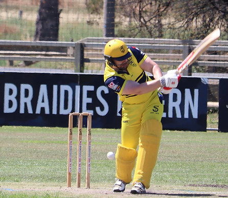 Cricket player in yellow uniform batting on the field with wickets behind and a sign that reads 'BRAIDIES.'