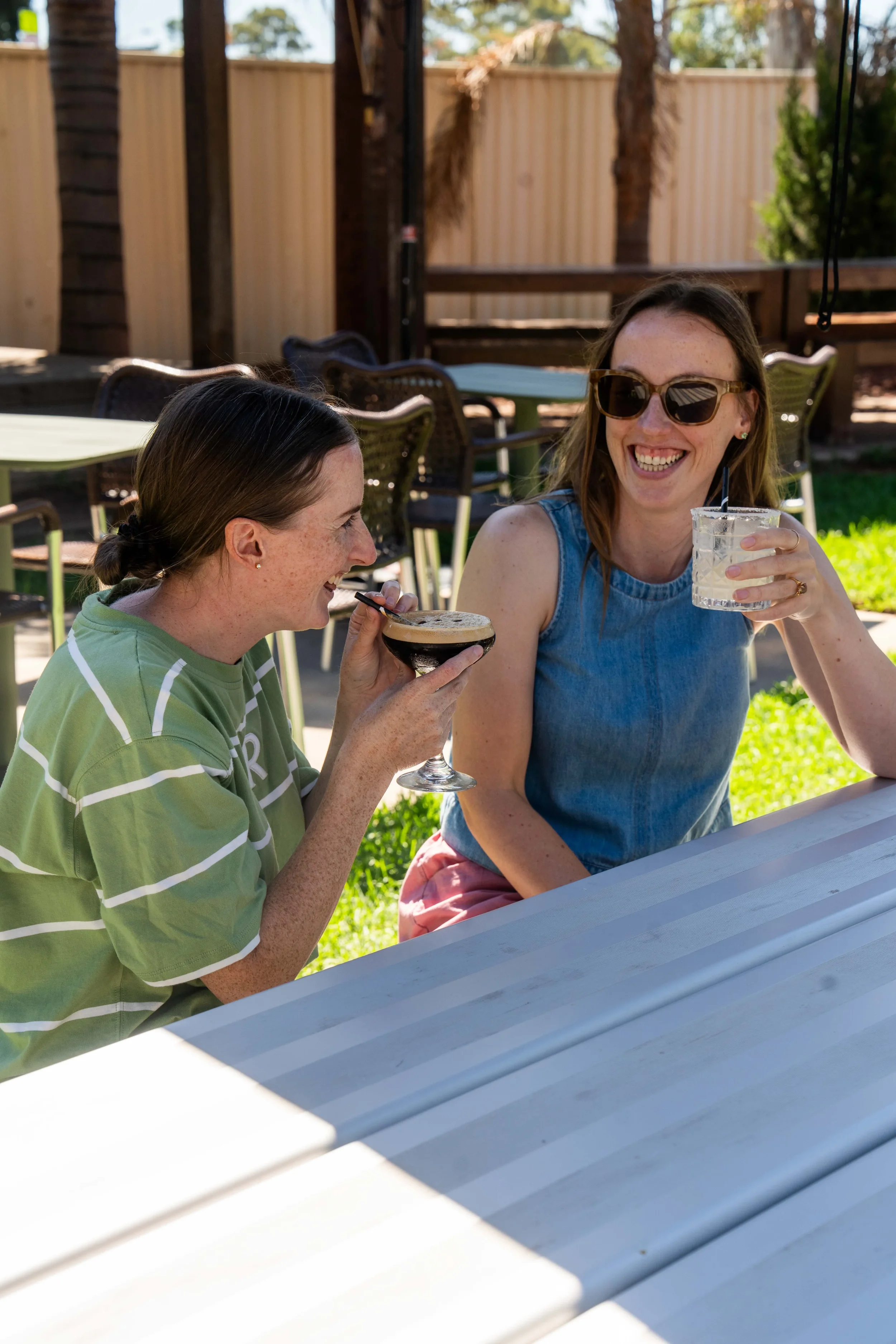 Two women sitting outdoors at a table, laughing and smiling. One is holding a cocktail glass, and the other has a glass with a clear drink and a straw. They are in a backyard with a wooden fence, plants, and trees.