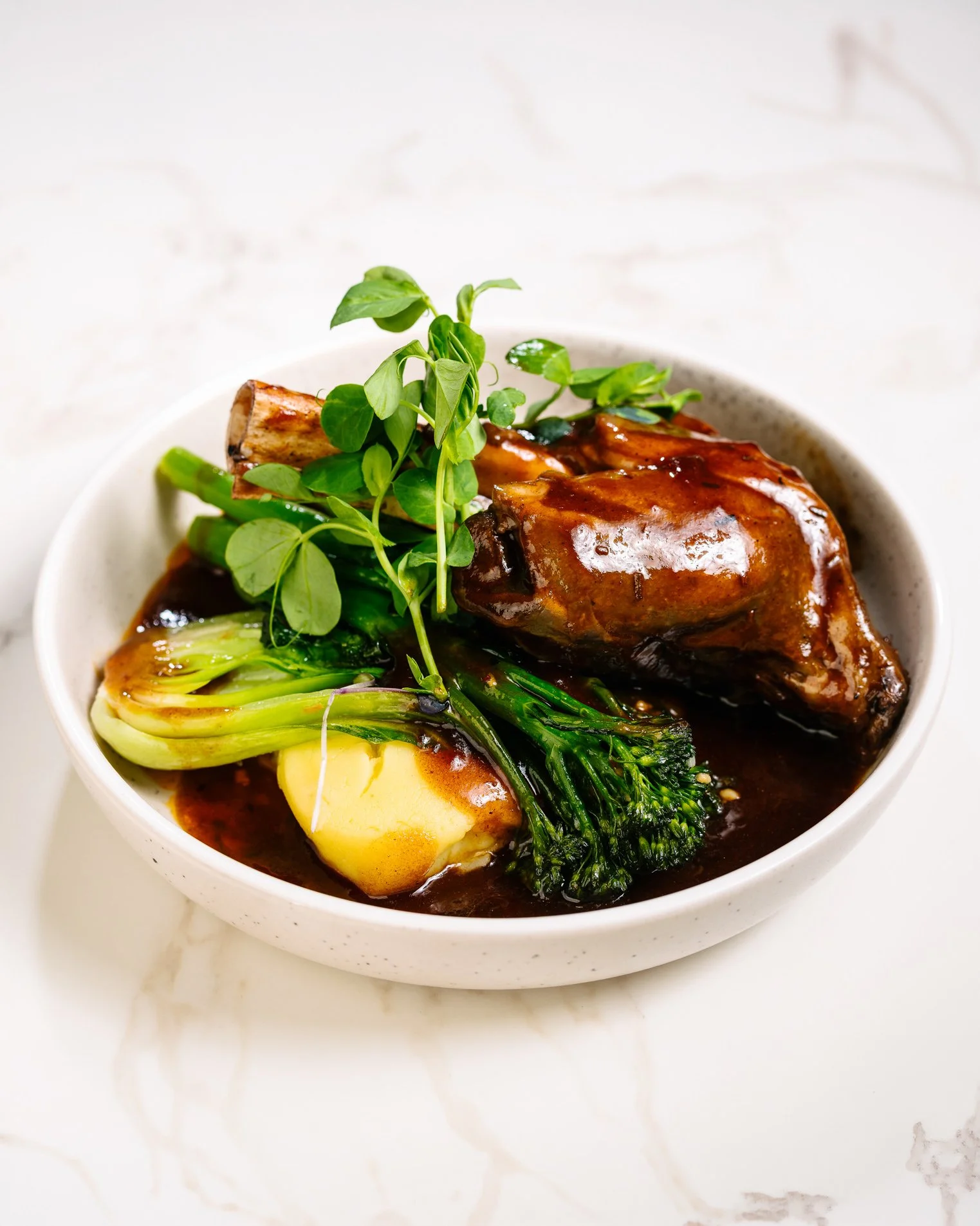 A bowl of beef stew with vegetables including broccoli, green onions, and a boiled egg, garnished with fresh herbs, in a white bowl on a white marble surface.