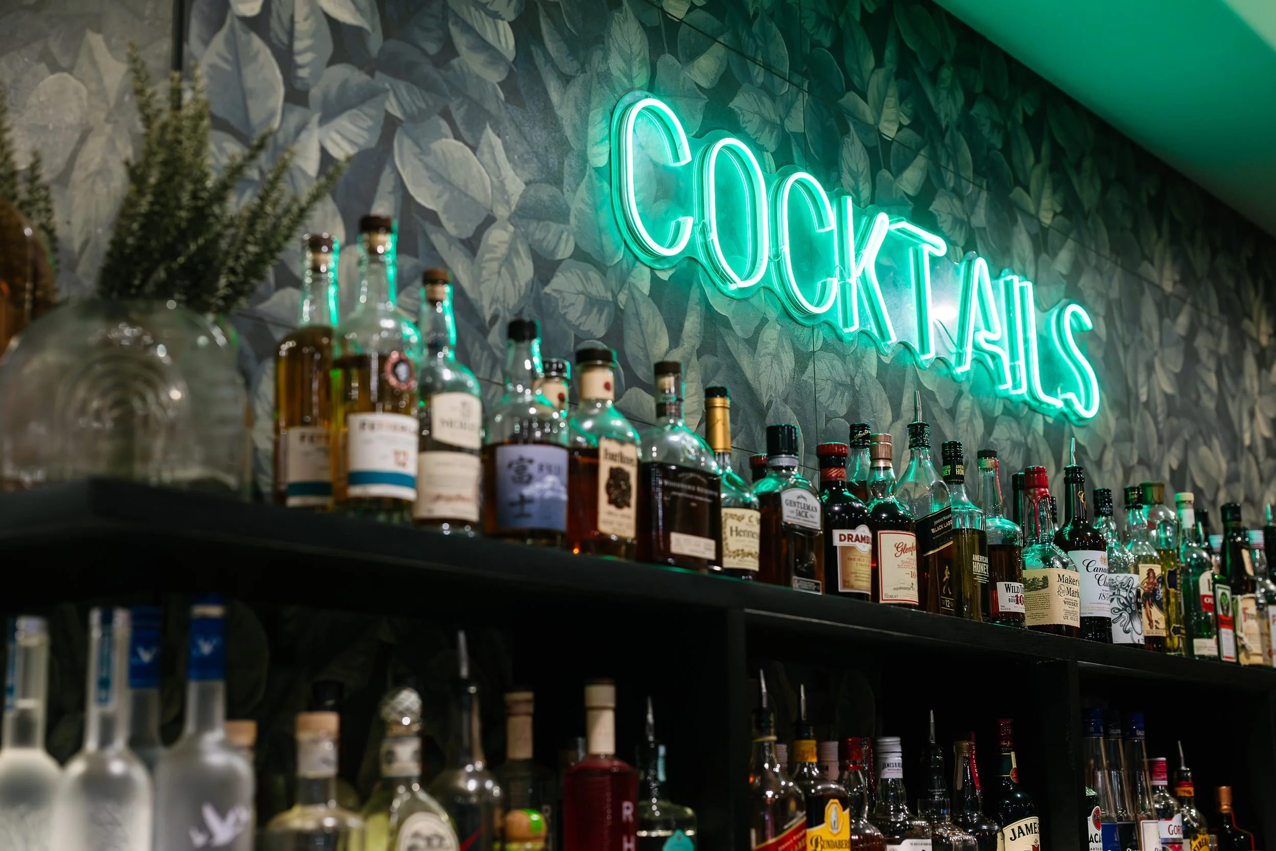 A neon sign that says 'cocktails' hangs on a wall decorated with large green leafy patterns. Below the sign, shelves display various bottles of liquor and mixers.