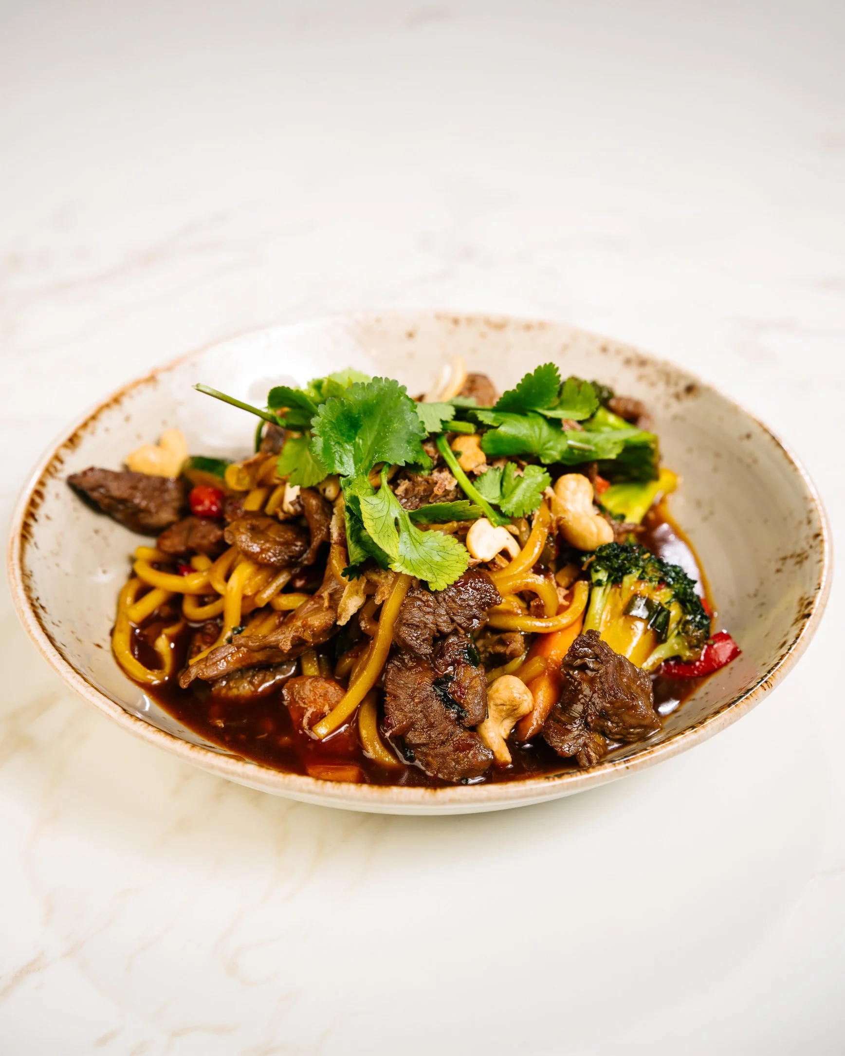 A bowl of Asian beef noodle stir-fry garnished with cilantro on a white surface.