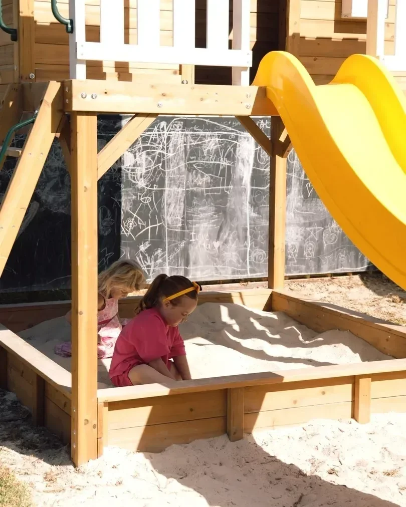 Two young girls playing in a sandbox at a playground, with a wooden play structure and yellow slide behind them, and a black chalkboard wall with scribbles in the background.