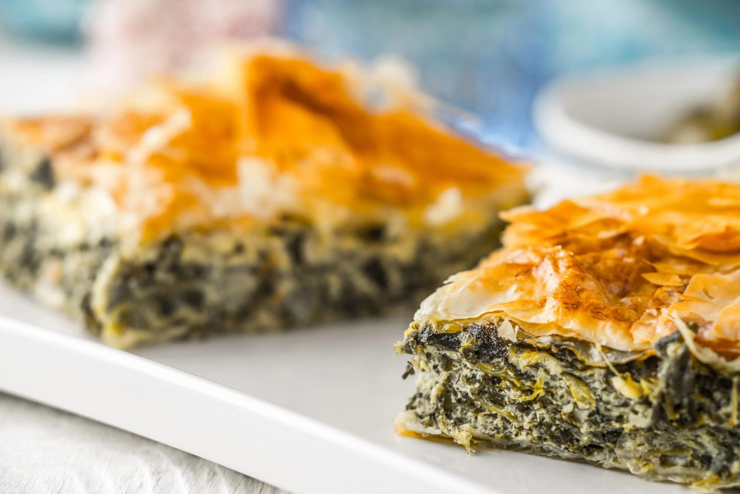 Close-up of a slice of savory pie with a flaky golden crust, filled with spinach and cheese, on a white plate.