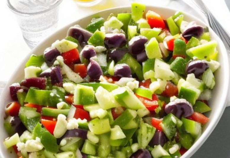 Bowl of chopped vegetables including celery, bell peppers, olives, tomatoes, and cucumbers, with feta cheese crumbles, next to a glass of water.