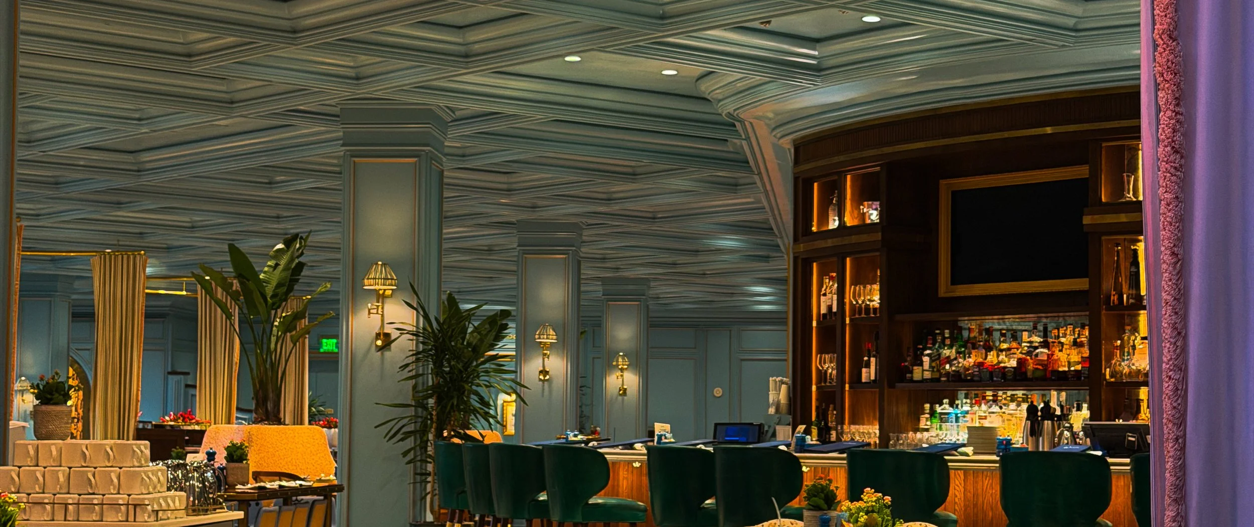 Interior of a restaurant or bar with a curved wooden bar counter, shelves with bottles and glassware, and mood lighting. There are green chairs, plants, and decorative lamps on the walls.